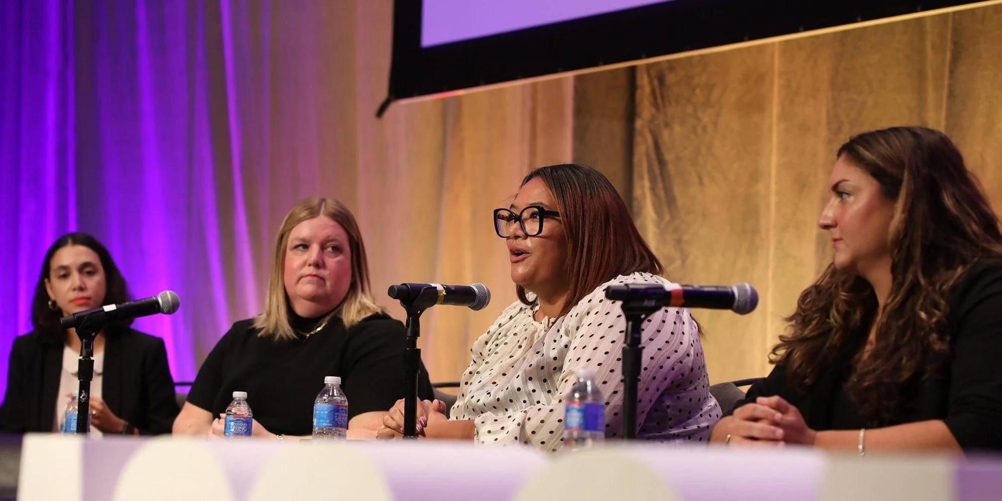 Four women, one of whom is Jenn Patrice Schultz, are seated at a panel discussion with microphones and water bottles on a table. The woman second from the right is speaking. The background is purple and gold.