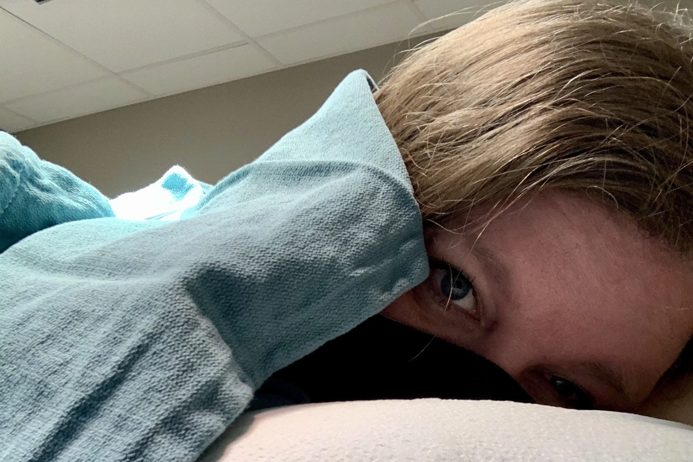 Jenn Patrice Schultz, a young woman with dark blonde hair lying on a surgical bedd, with their face partially visible, covered in a light blue medical blanket.