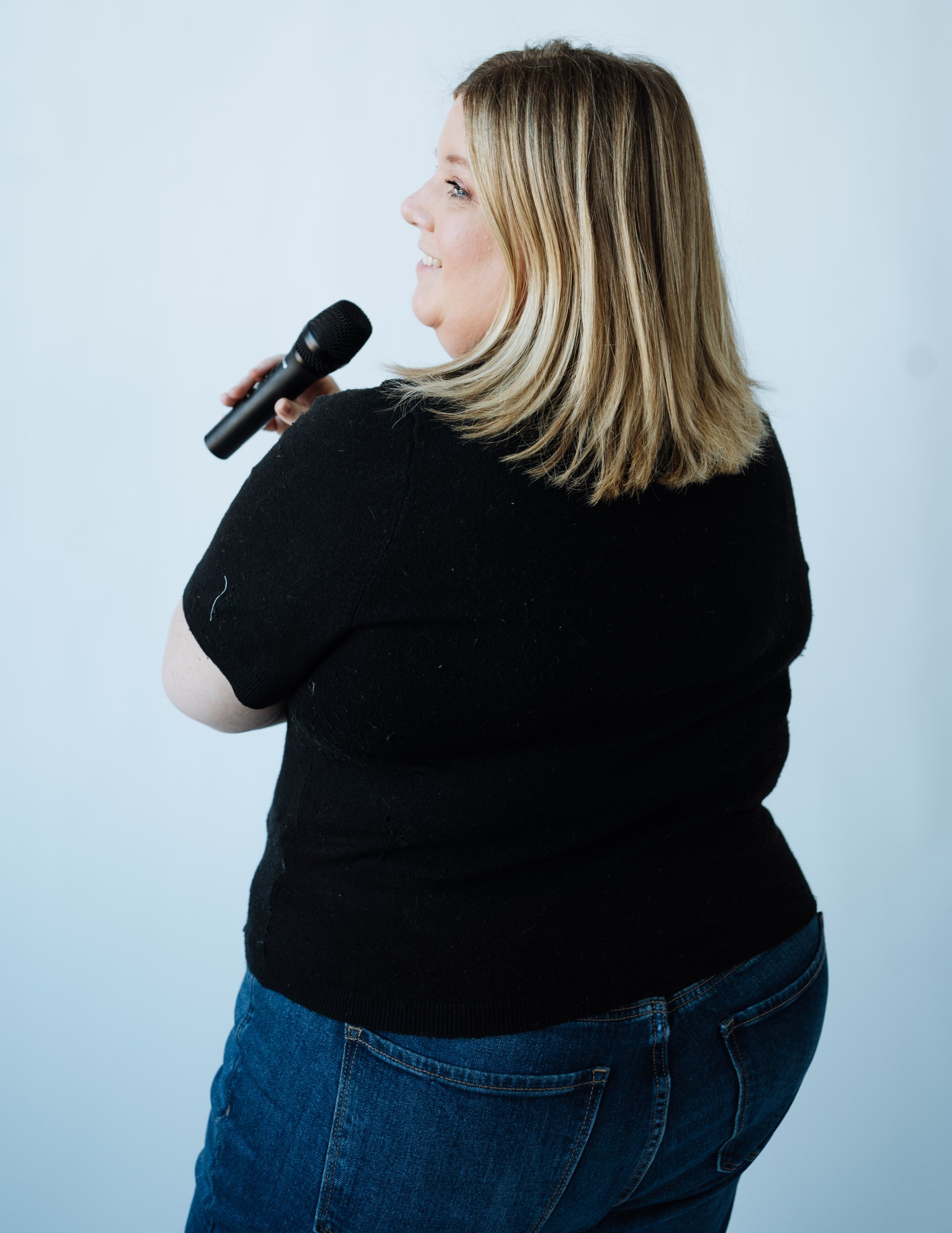 Jenn Patrice Schultz, a woman with shoulder-length blonde hair, wearing a black shirt and blue jeans, holding a microphone and smiling, standing against a plain light-colored background.