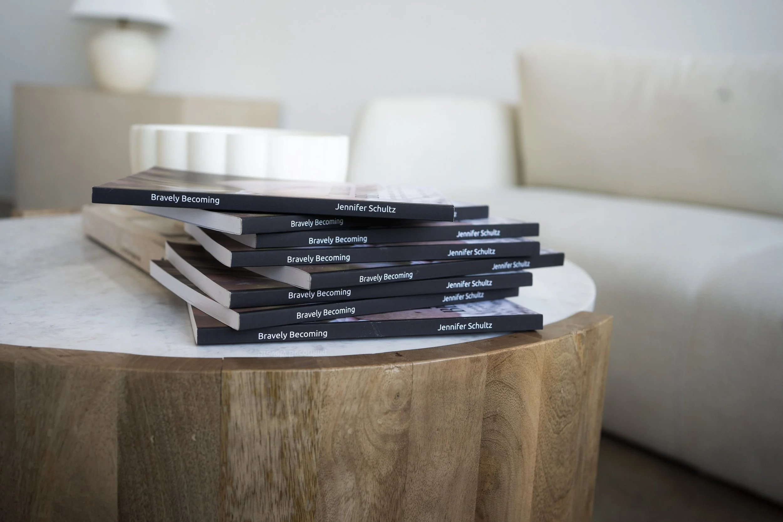 A stack of books titled 'Bravely Becoming' by Jennifer Schultz on a round wooden table.