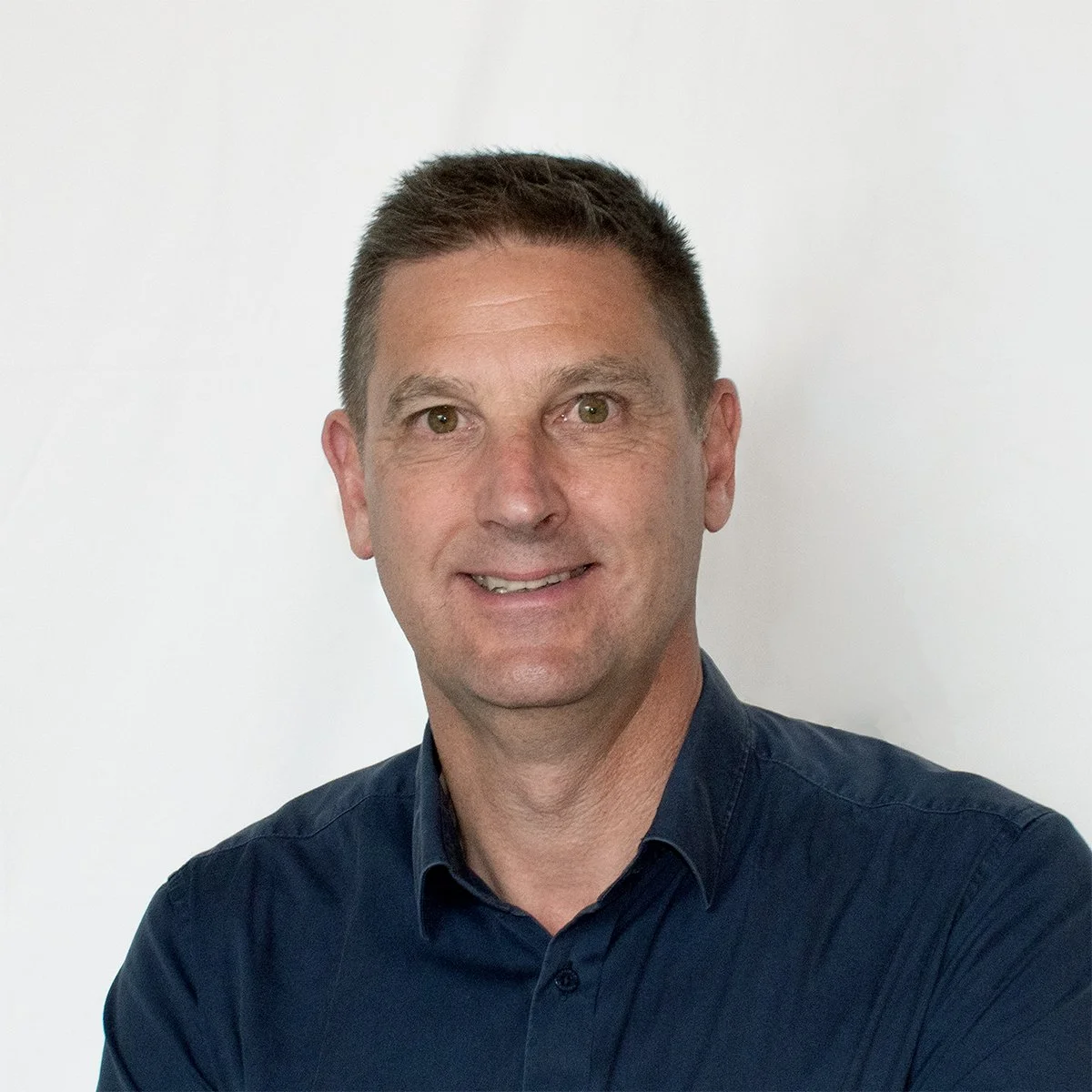 Portrait of a man with short brown hair, wearing a dark blue button-up shirt, smiling, against a plain white background.