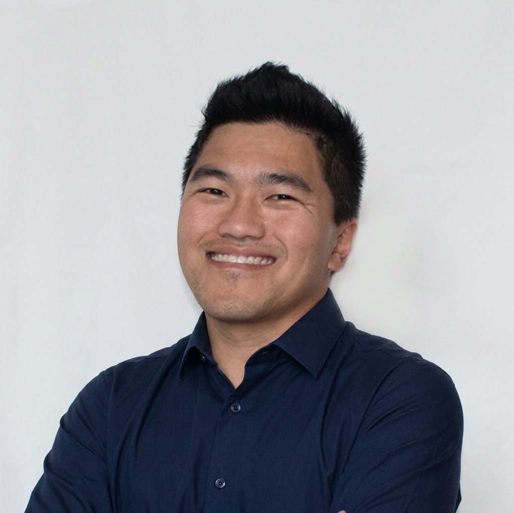 A smiling man with short black hair, wearing a dark button-up shirt, standing against a plain white background.
