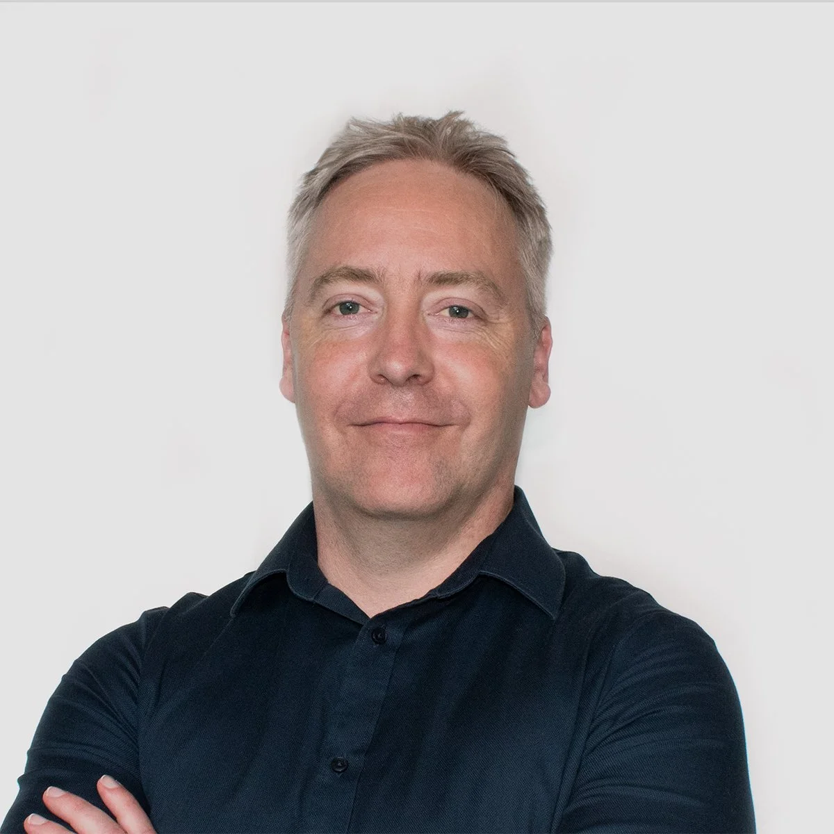 Portrait of a middle-aged man with short, gray hair wearing a black collared shirt, standing against a plain white background.