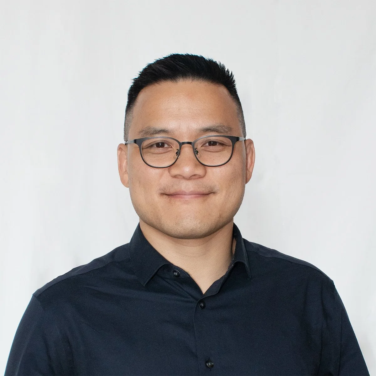 Portrait of a man wearing glasses and a dark button-up shirt, smiling, standing against a plain white background.
