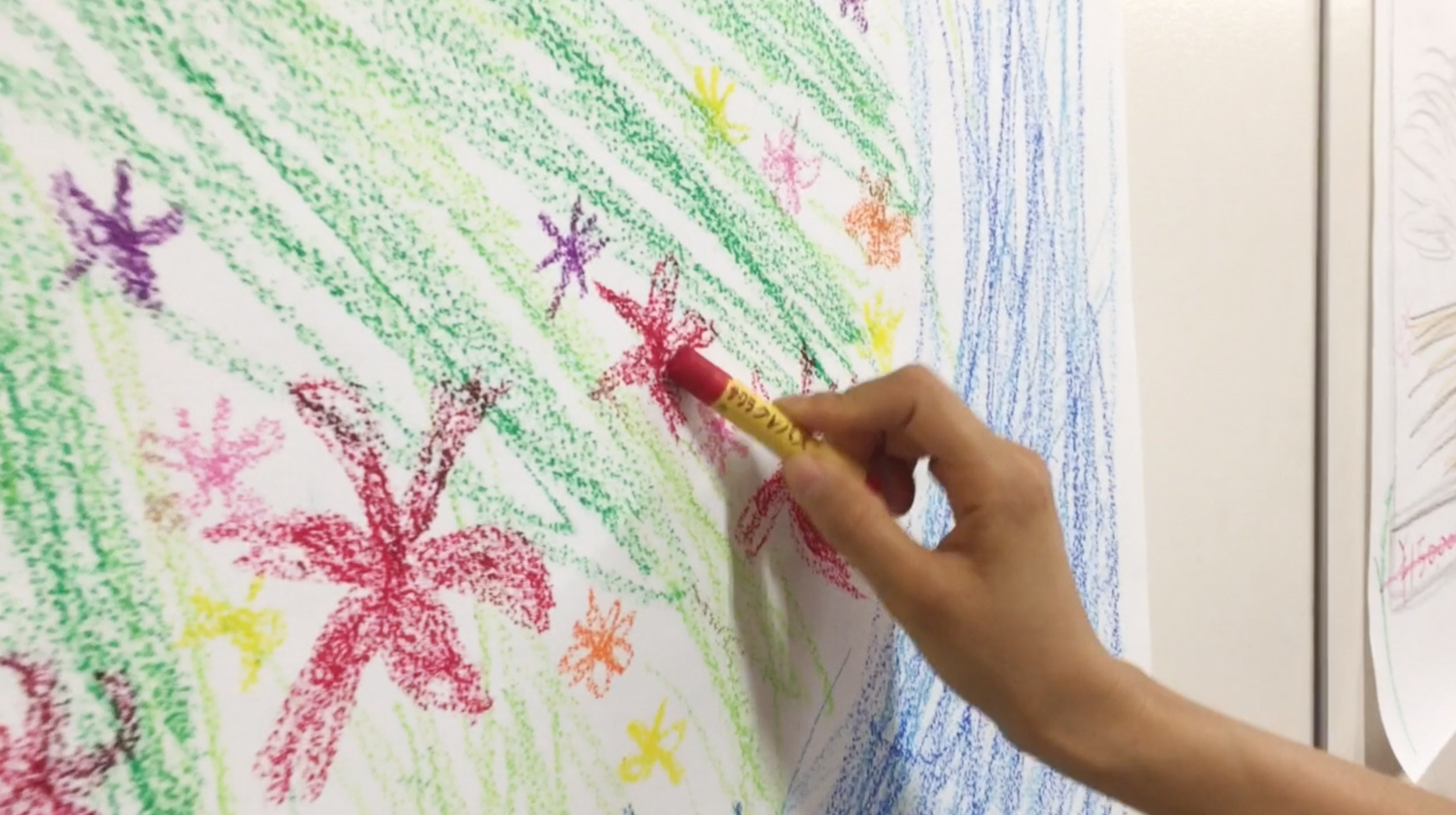 Child's colorful drawing of flowers, trees, and plants being colored with crayons by a person's hand.