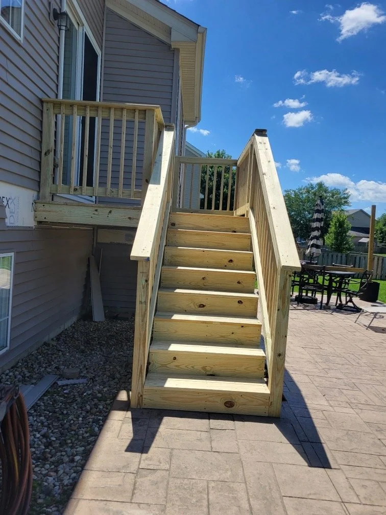 New wooden outdoor staircase leading to a second-story deck with a railing, attached to a house with vinyl siding, in a backyard with a paved patio and outdoor furniture.