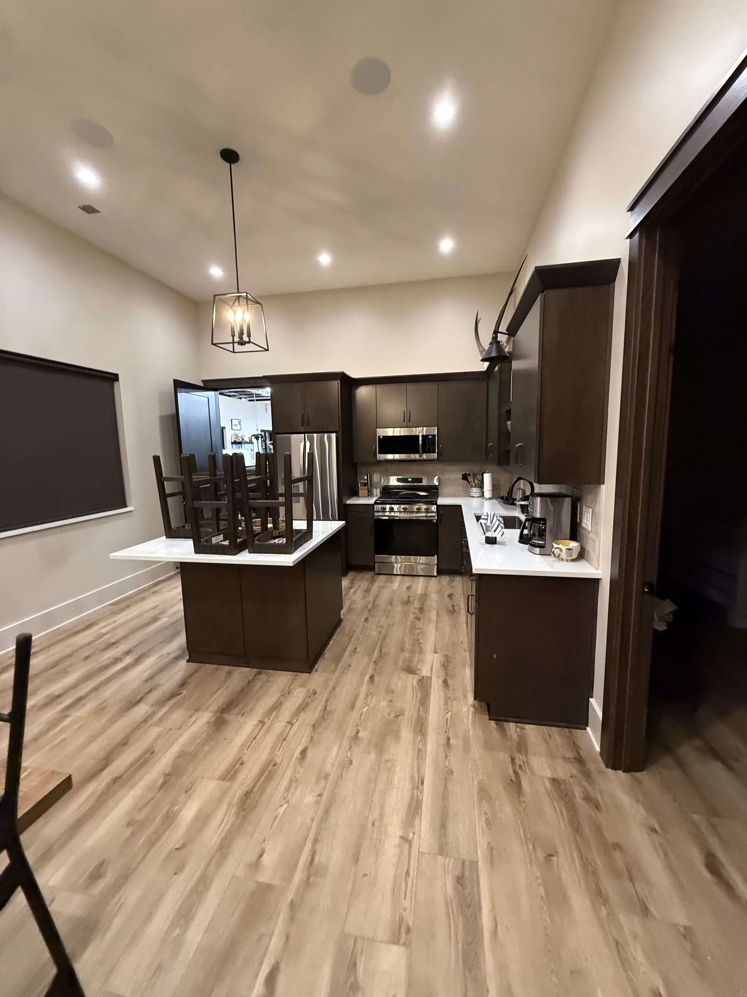 Empty kitchen with dark wood cabinets, stainless steel appliances, white countertops, and wooden flooring, with chairs placed upside down on the kitchen island.