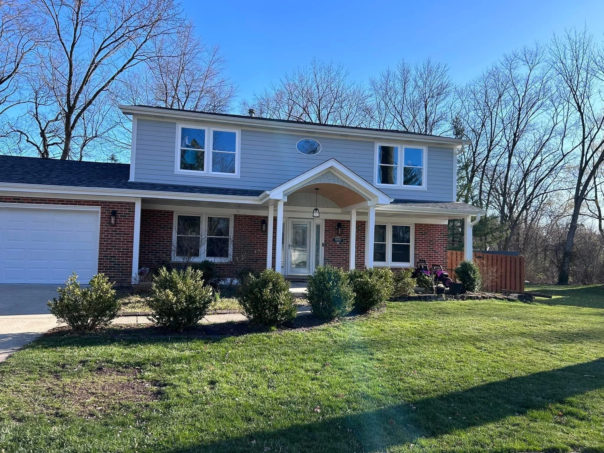 A two-story suburban house with a brick and siding exterior, front porch with columns, and a garage. Well-maintained lawn with bushes and a small garden.
