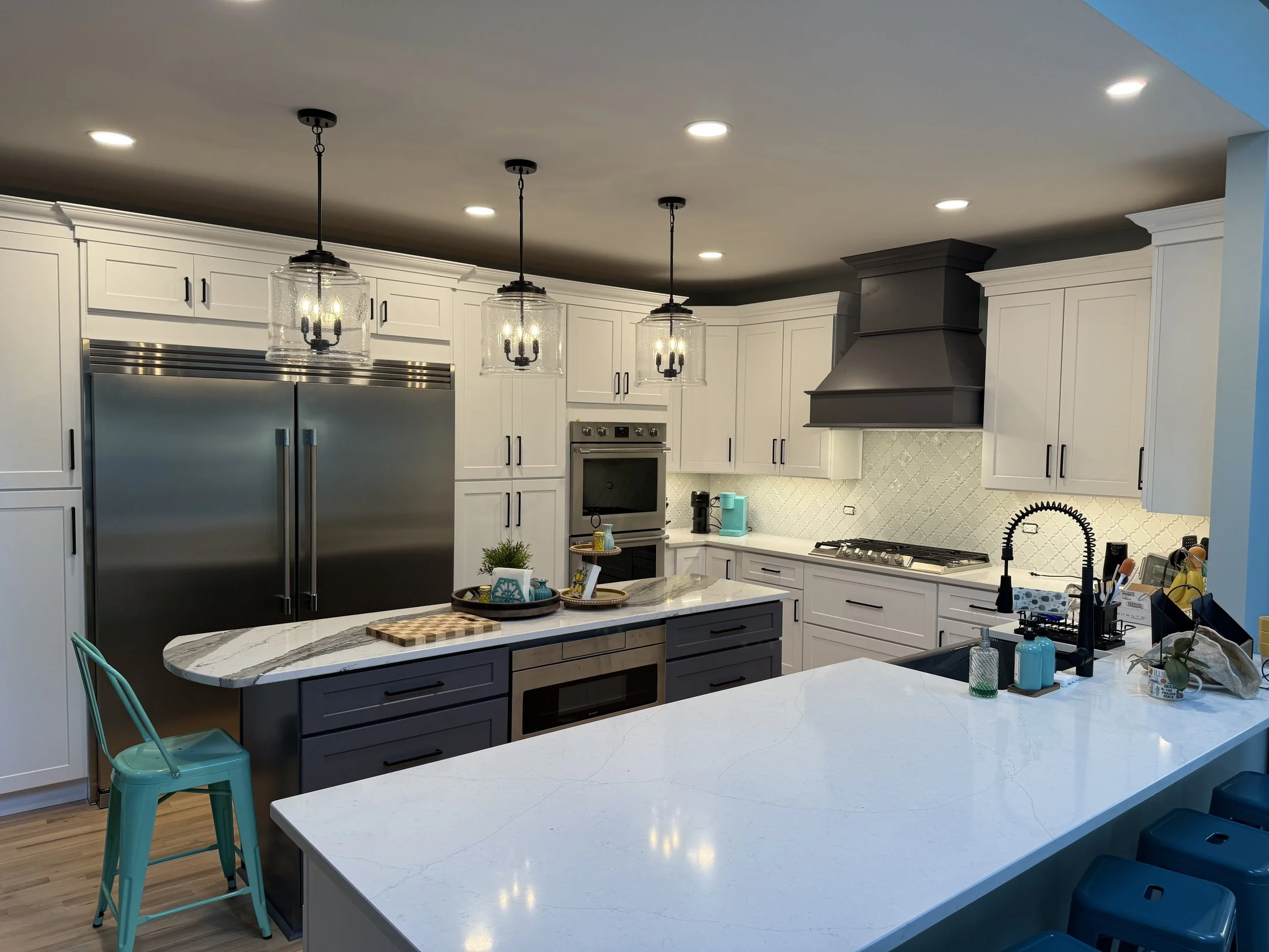 Modern kitchen with white cabinets, stainless steel refrigerator, black range hood, white marble countertops, and a kitchen island with dark gray cabinets. There are three pendant lights above the island and recessed ceiling lights. Various kitchen appliances and decor items are visible.