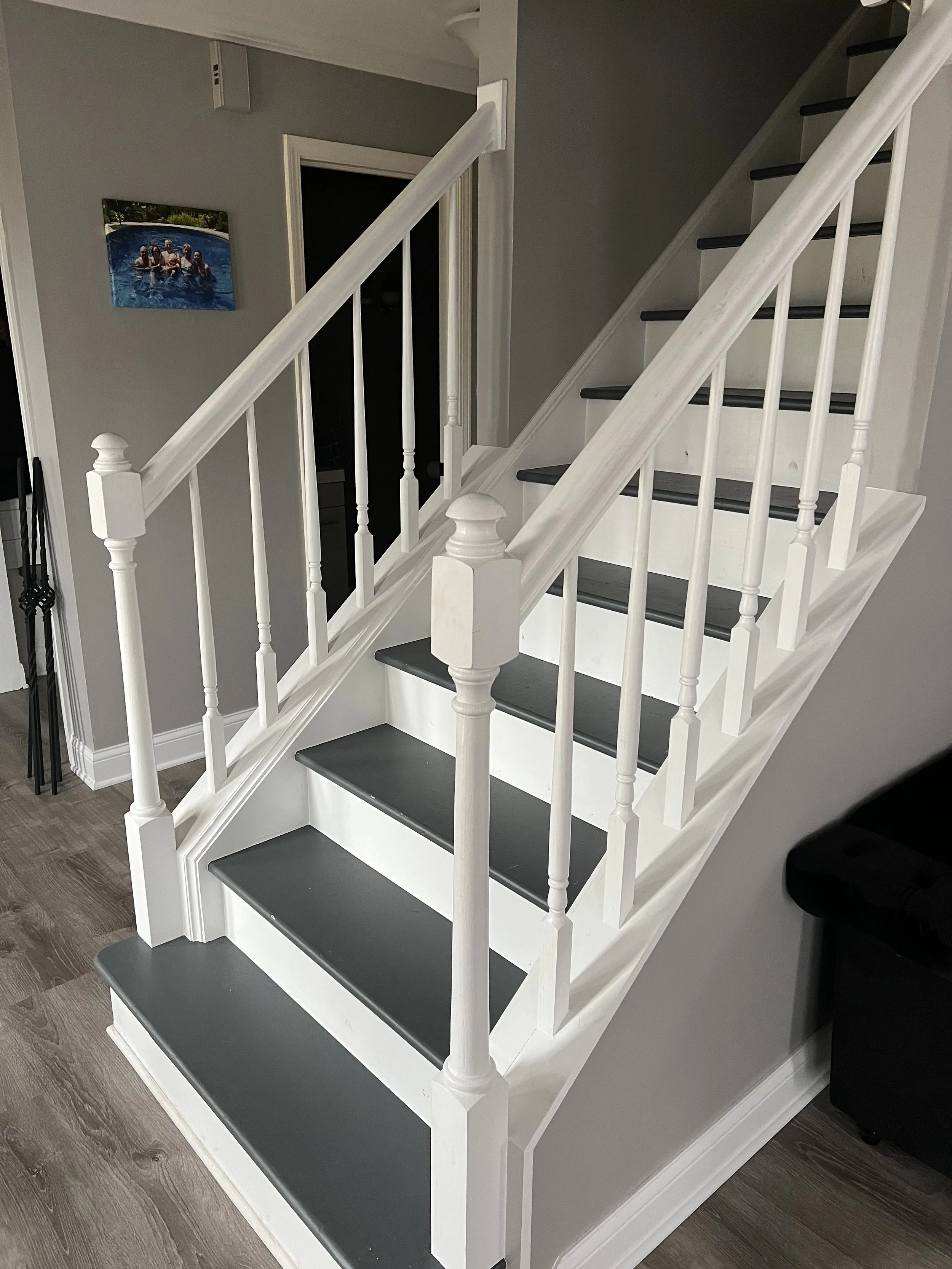 Interior view of a staircase with white railings and dark gray steps, positioned next to a gray wall. There is a framed photo of people in a swimming pool on the wall near the staircase.