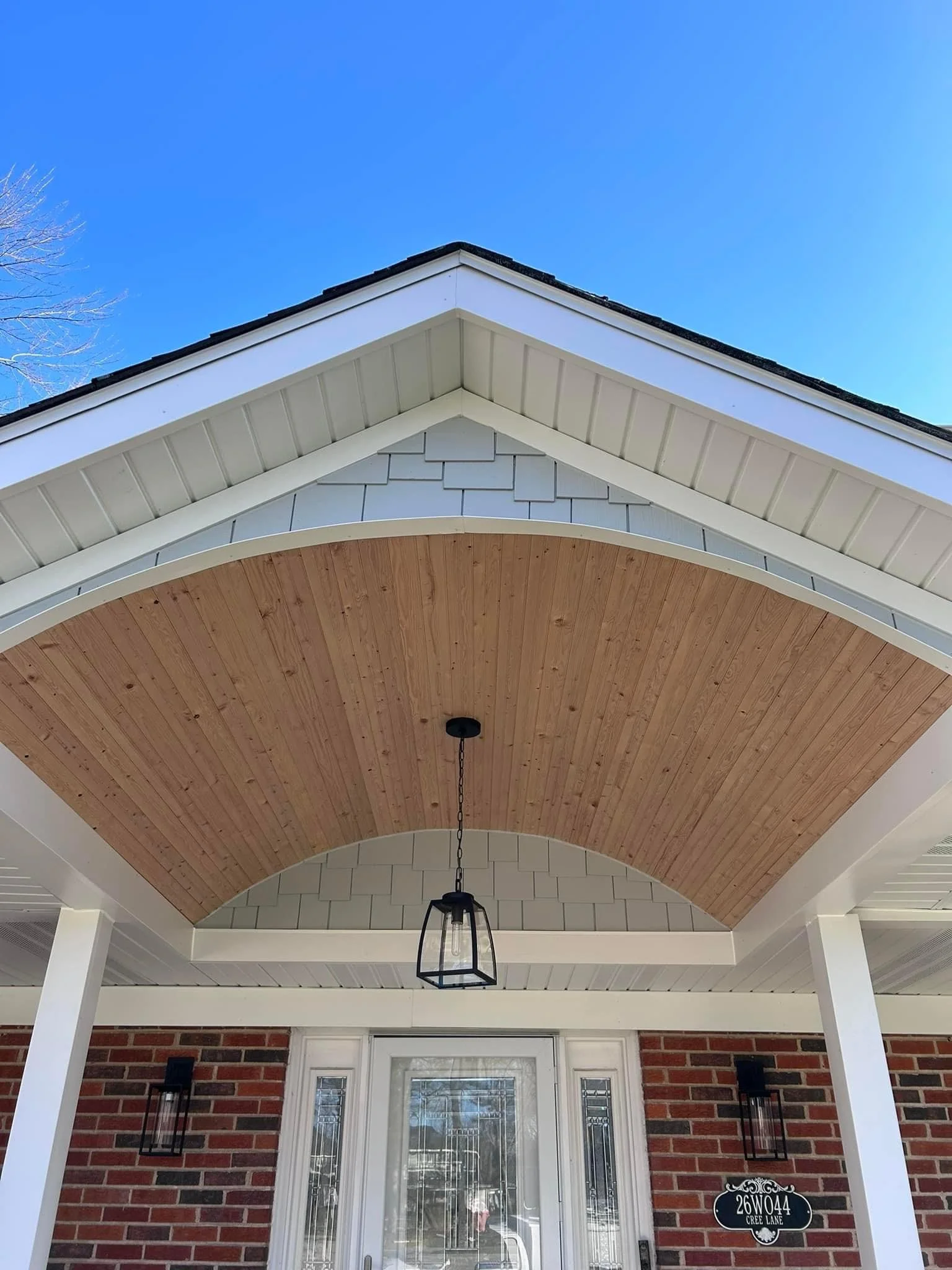 Front porch of a house with a wooden ceiling, brick walls, a hanging light fixture, and house number 26W044 on a black plaque.