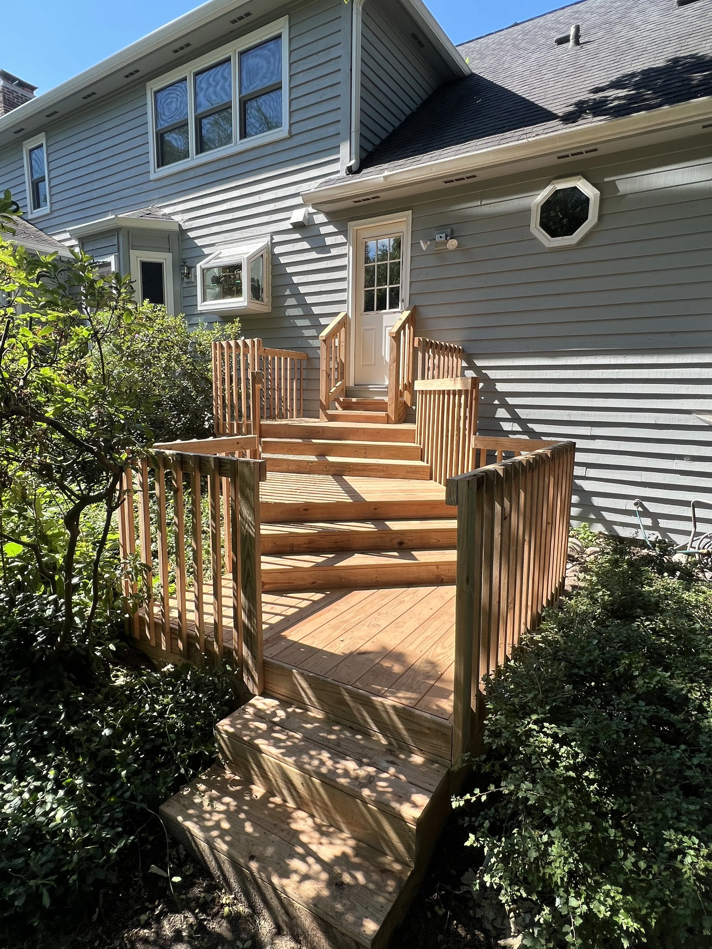 Wooden deck with stairs leading up to a gray house with siding, multiple windows, some open, and a door. The deck is surrounded by greenery and shrubs, with shadows cast by nearby trees.