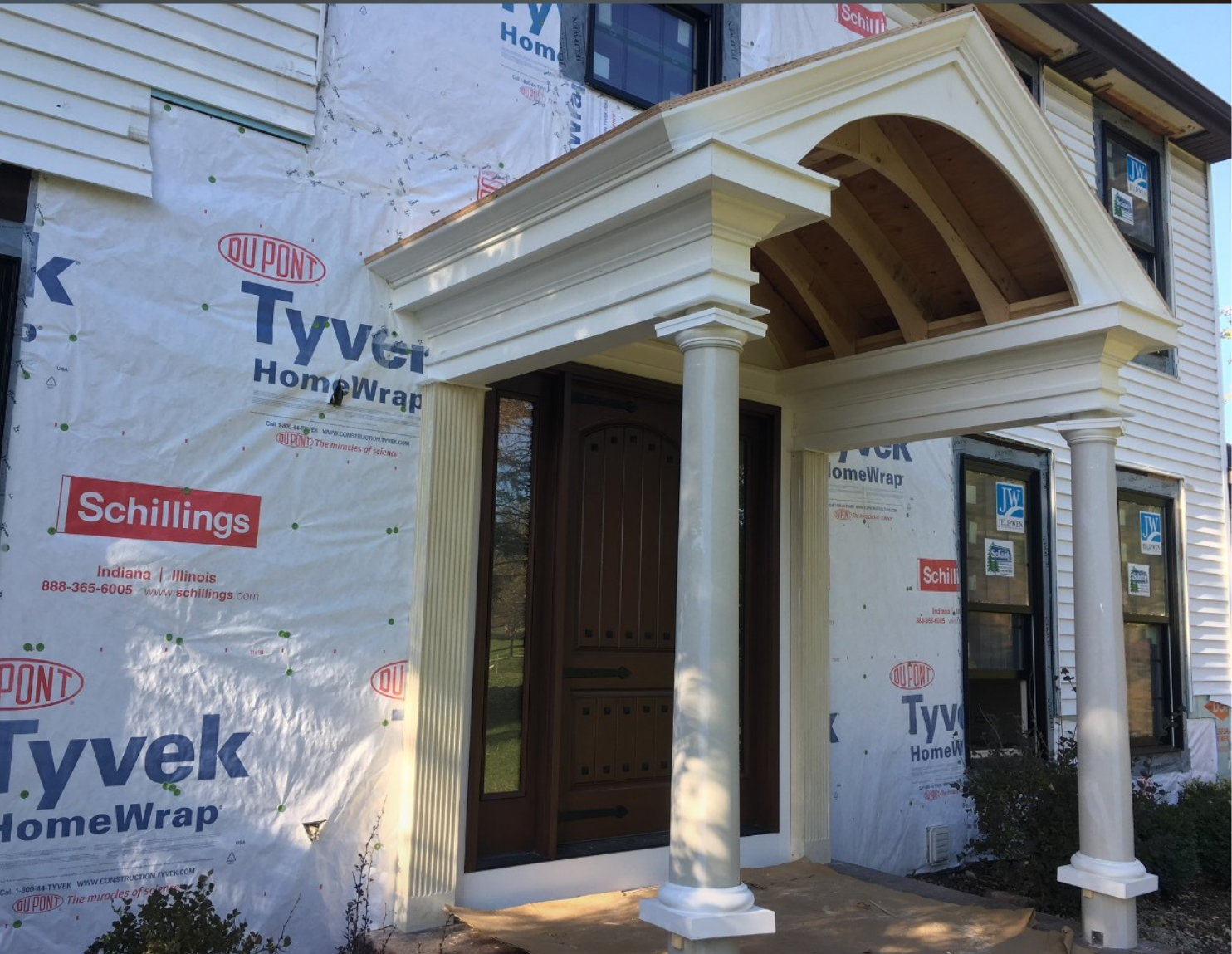 Under construction house with an ornate white porch and columns, beige front door, and windows, partially covered with Tyvek house wrap branded material.