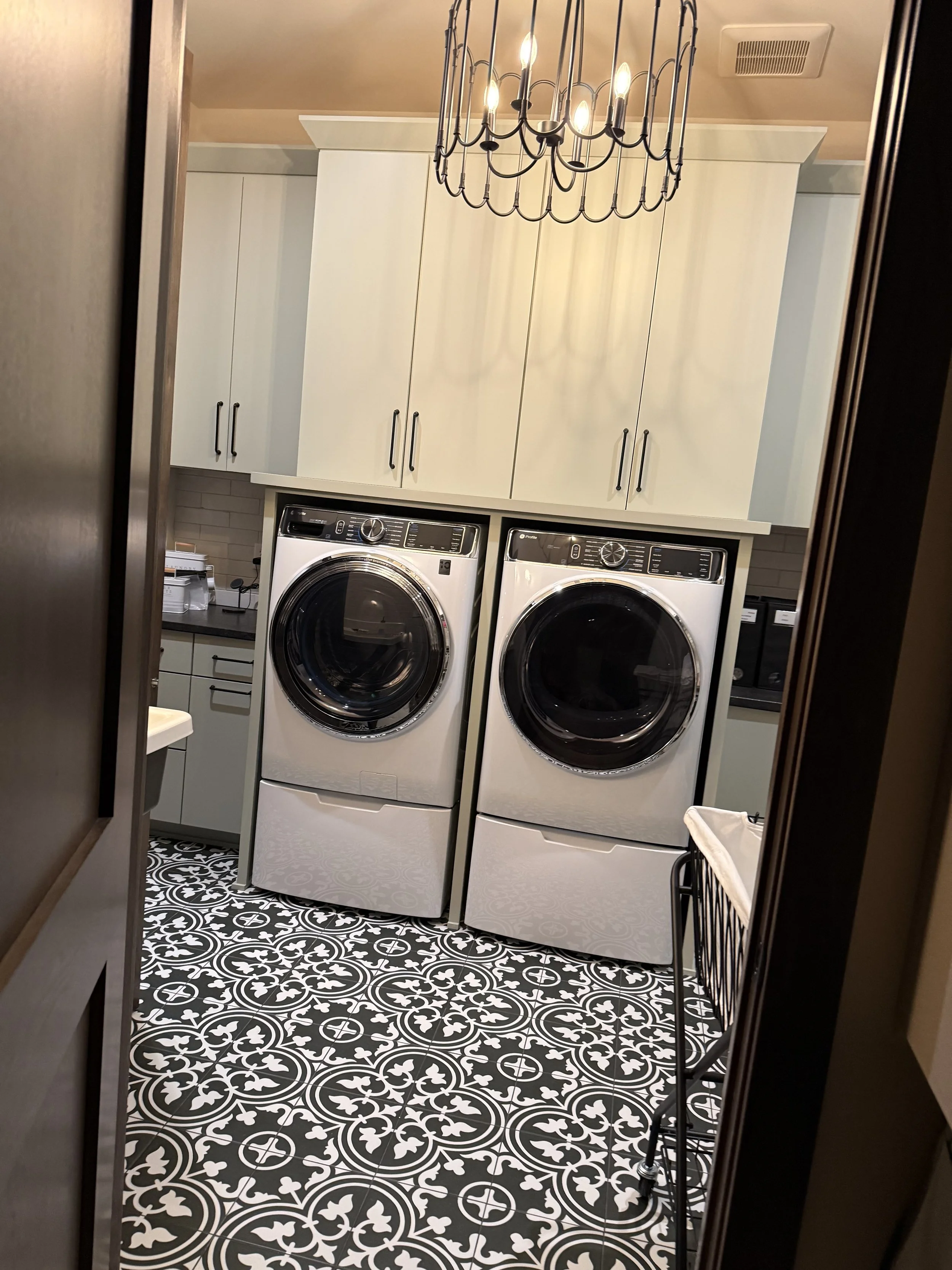 Laundry room with front-loading washer and dryer below white cabinets, black and white patterned tile floor, chandelier, and laundry baskets.