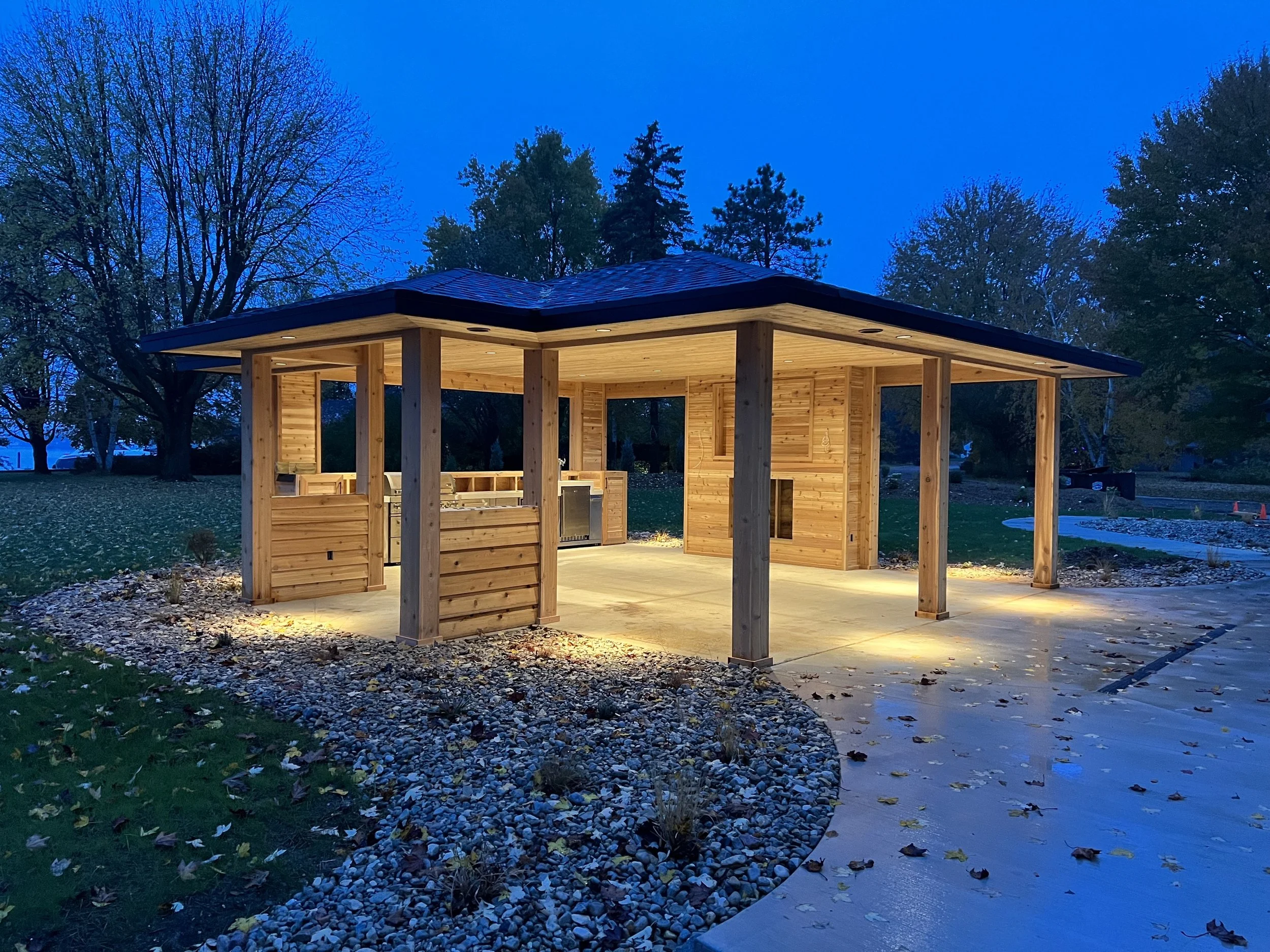 A wooden pavilion with a dark roof, illuminated by outdoor lights, situated in a park at twilight, surrounded by trees and a pebble-lined path.