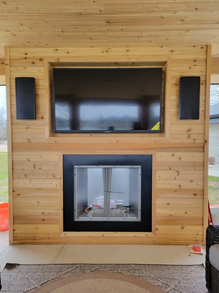 Wood-paneled wall with a mounted flat-screen TV above a modern fireplace with a glass front, surrounded by black trim, and two tall black speakers on either side.