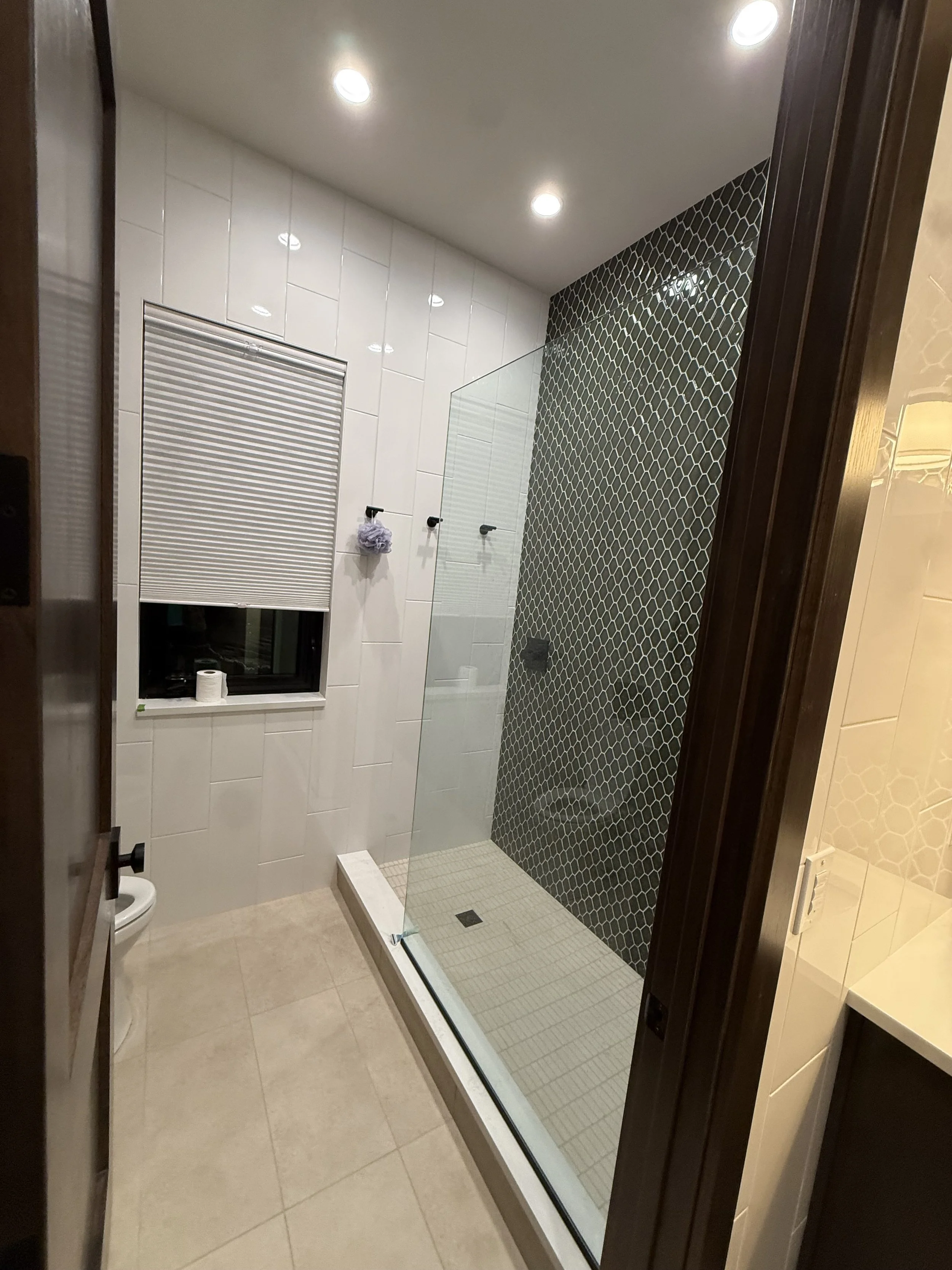 A modern bathroom with a glass shower enclosure, tiled walls, a window with a closed blind, a toilet, and recessed ceiling lights.