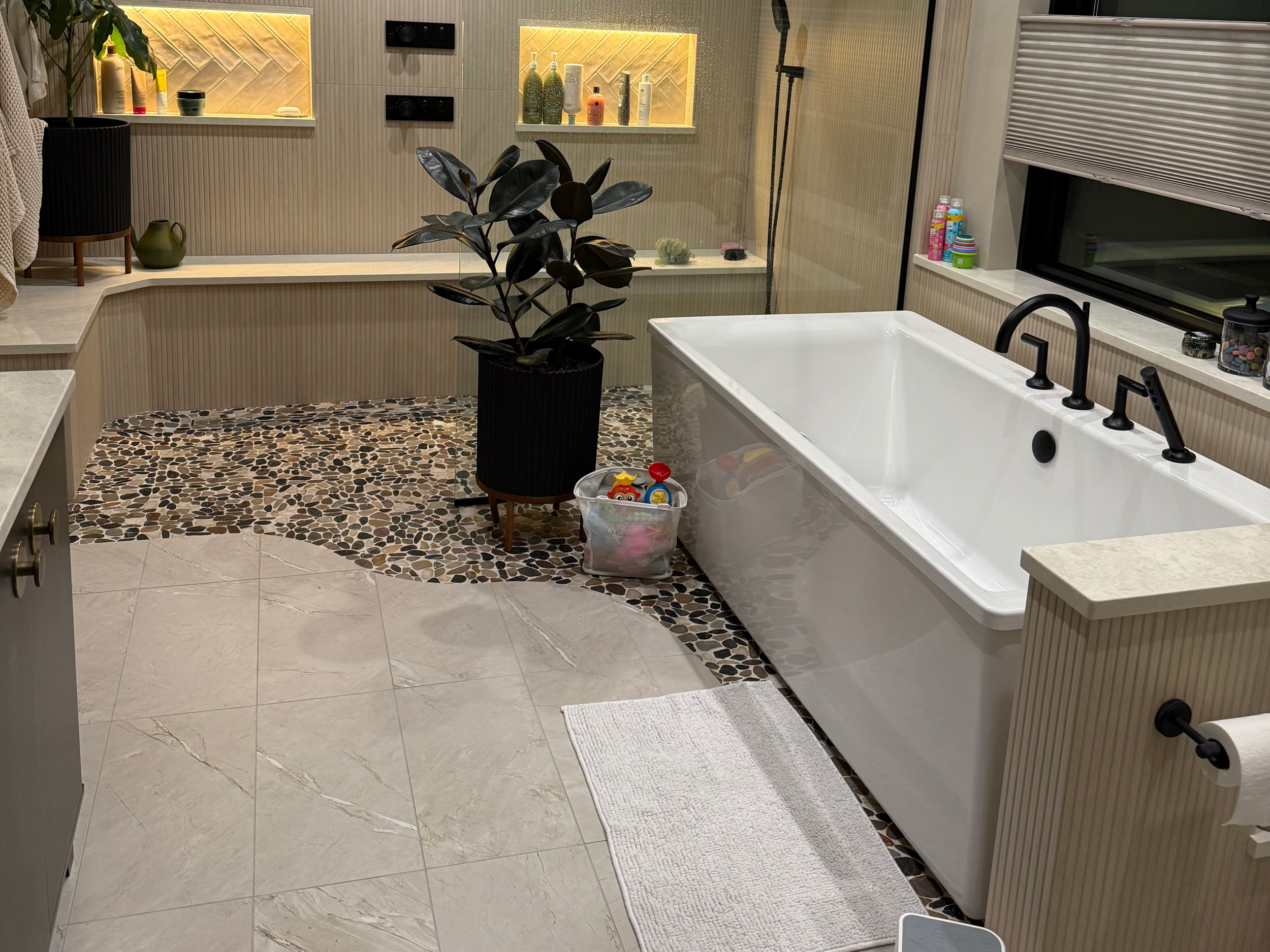 Modern bathroom with a large soaking tub, black fixtures, a mosaic pebble floor mat, a potted plant, and various toiletries on a windowsill.