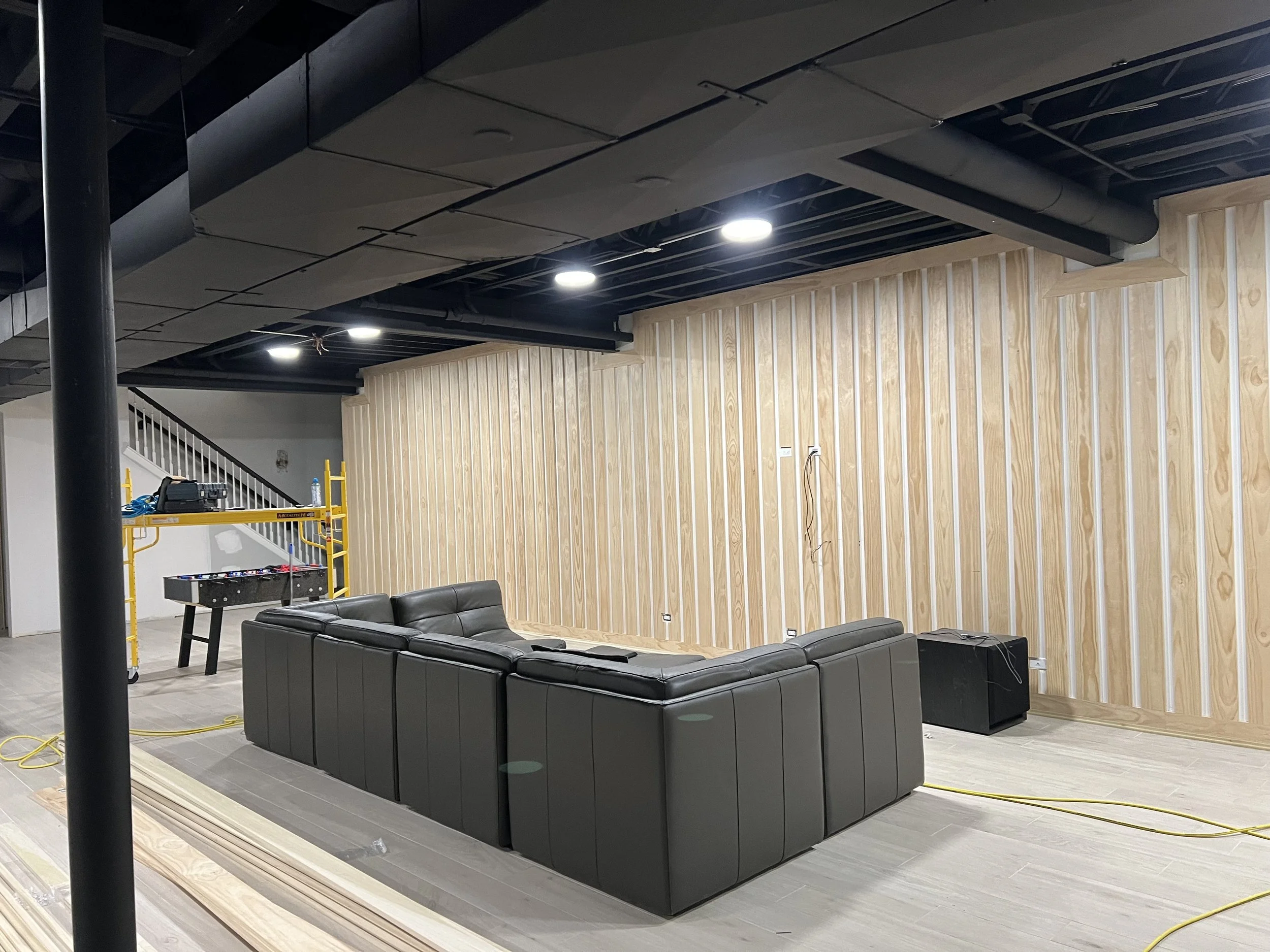 Interior of a room under construction with black sectional sofa, wooden wall paneling, construction tools, scaffolding, and yellow extension cords.