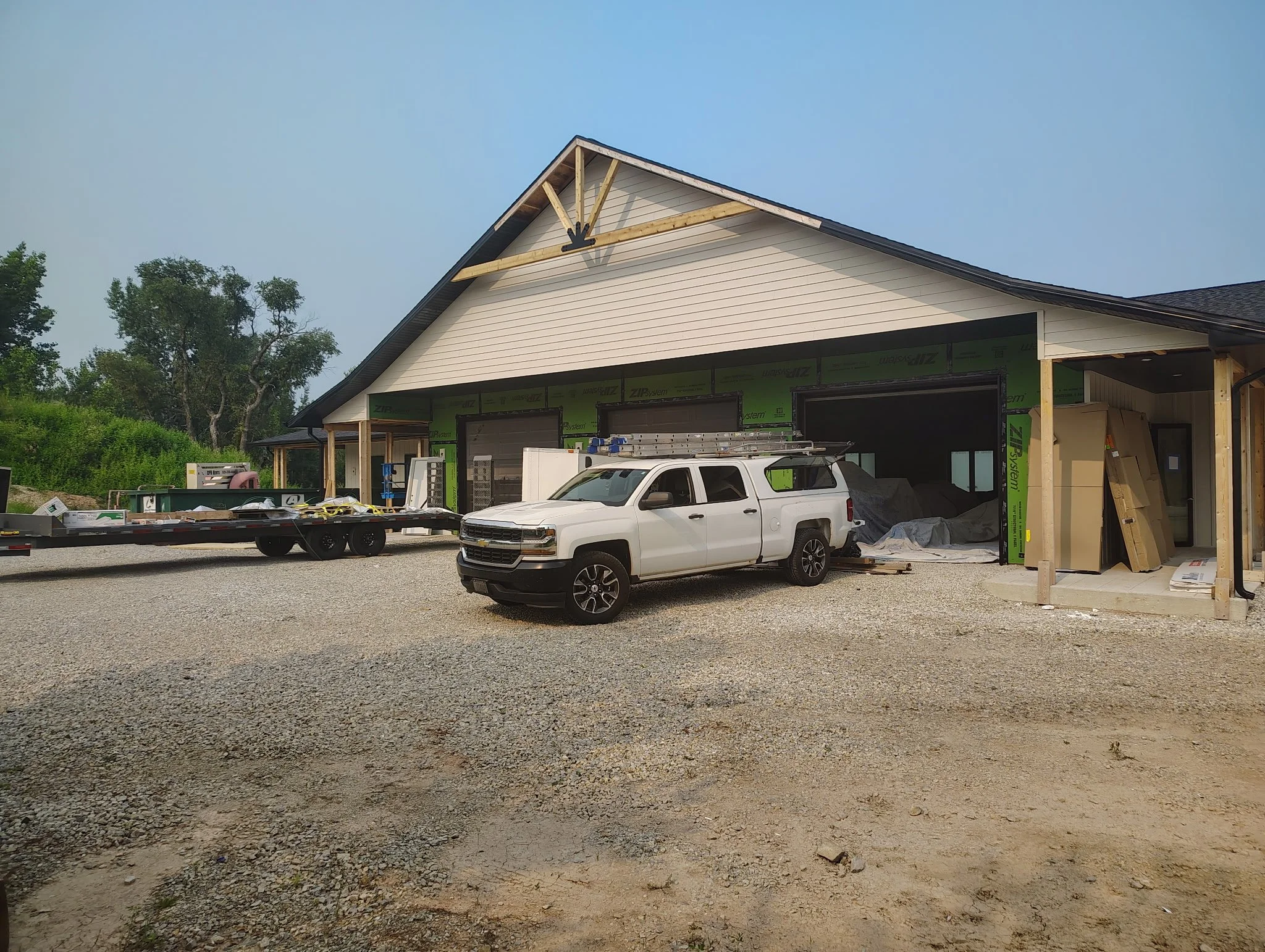 A house under construction with a white truck parked in front, construction materials and equipment around, and the house partially covered with building wrap.
