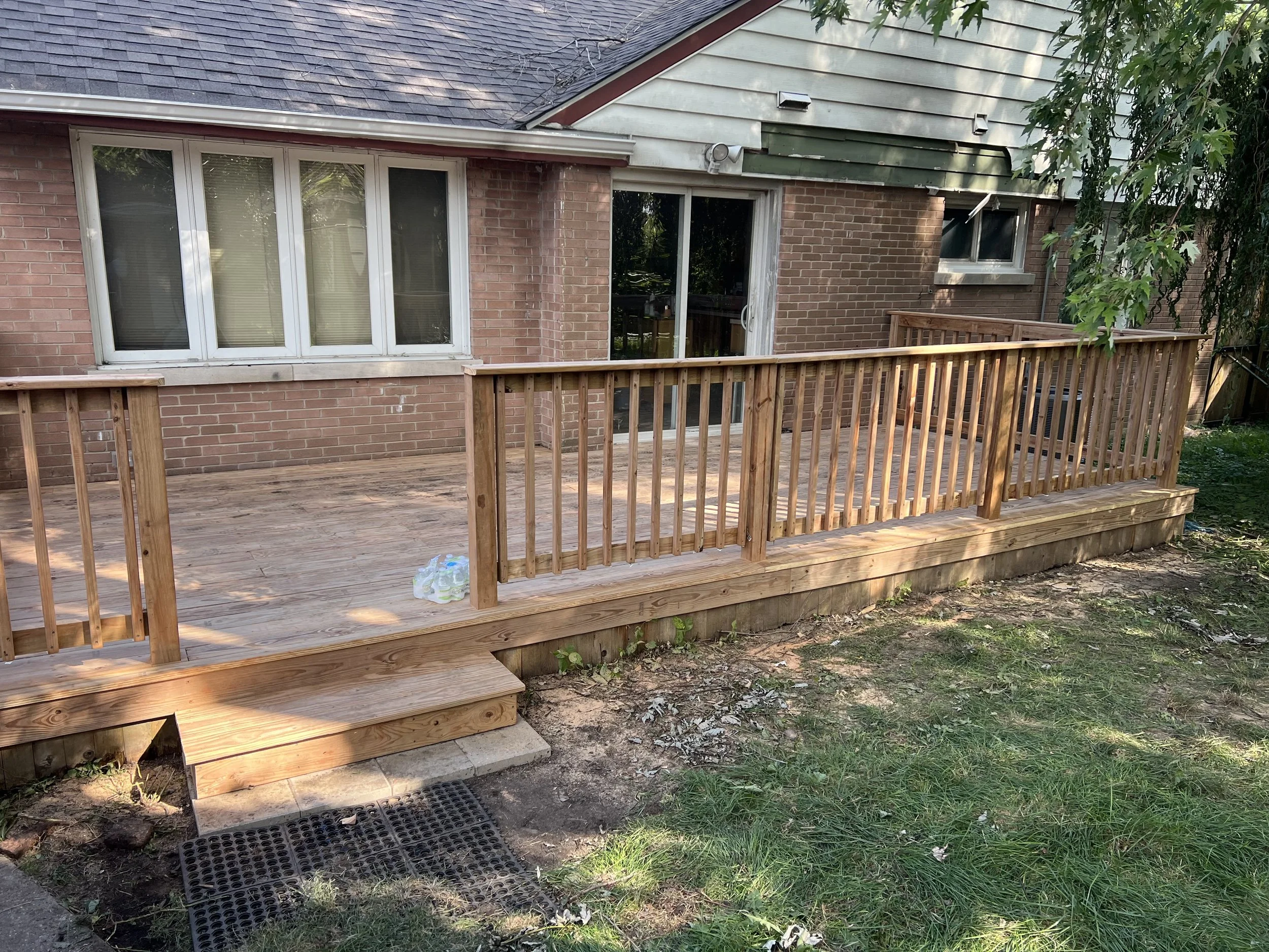 Newly built wooden deck with railing attached to the back of a brick house, with stairs leading down to a grassy yard.