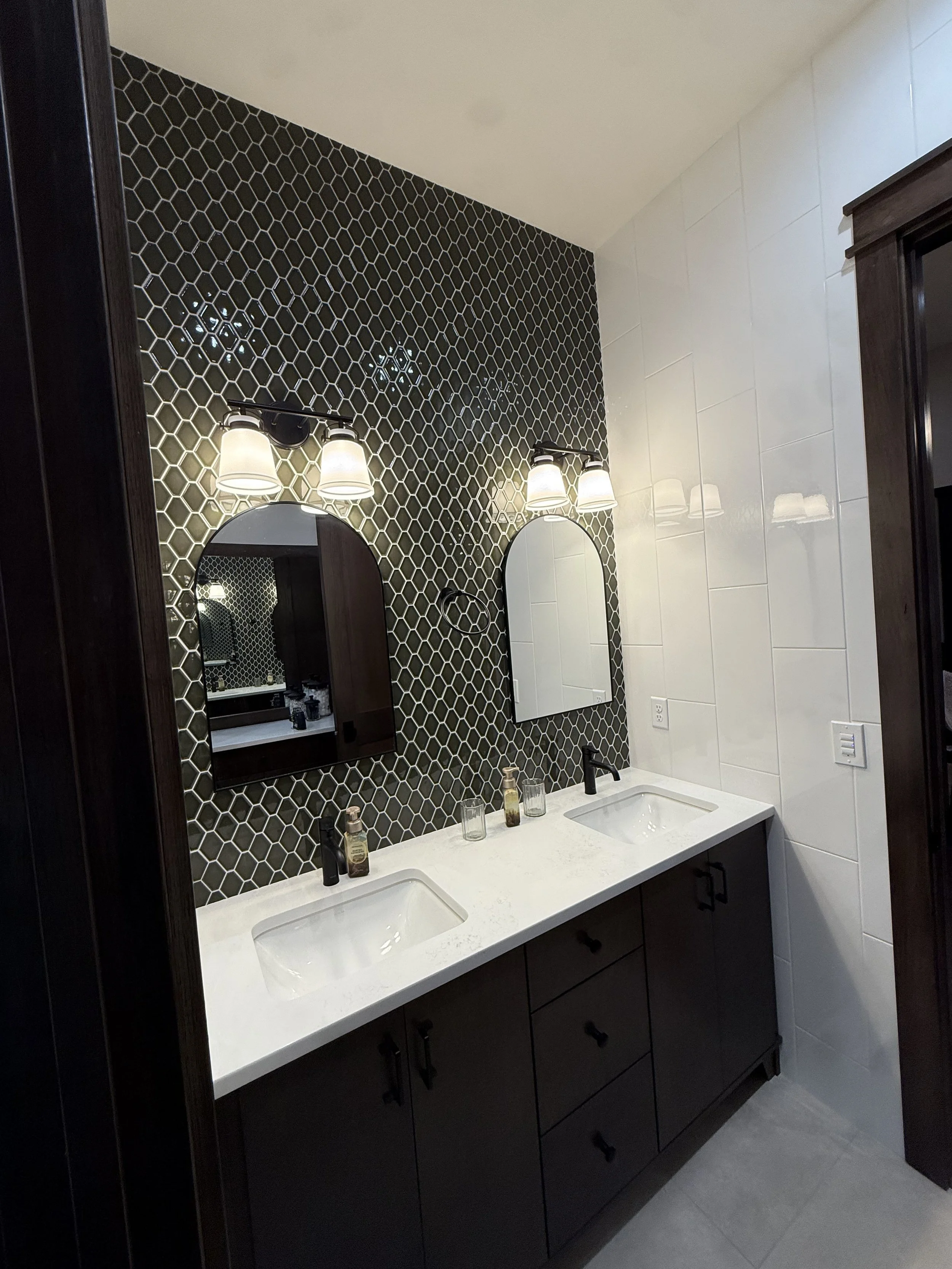Modern bathroom with a double vanity, black cabinets, white countertop, and two oval mirrors. The wall behind the mirrors has black honeycomb tiles, and there are two light fixtures above each mirror.
