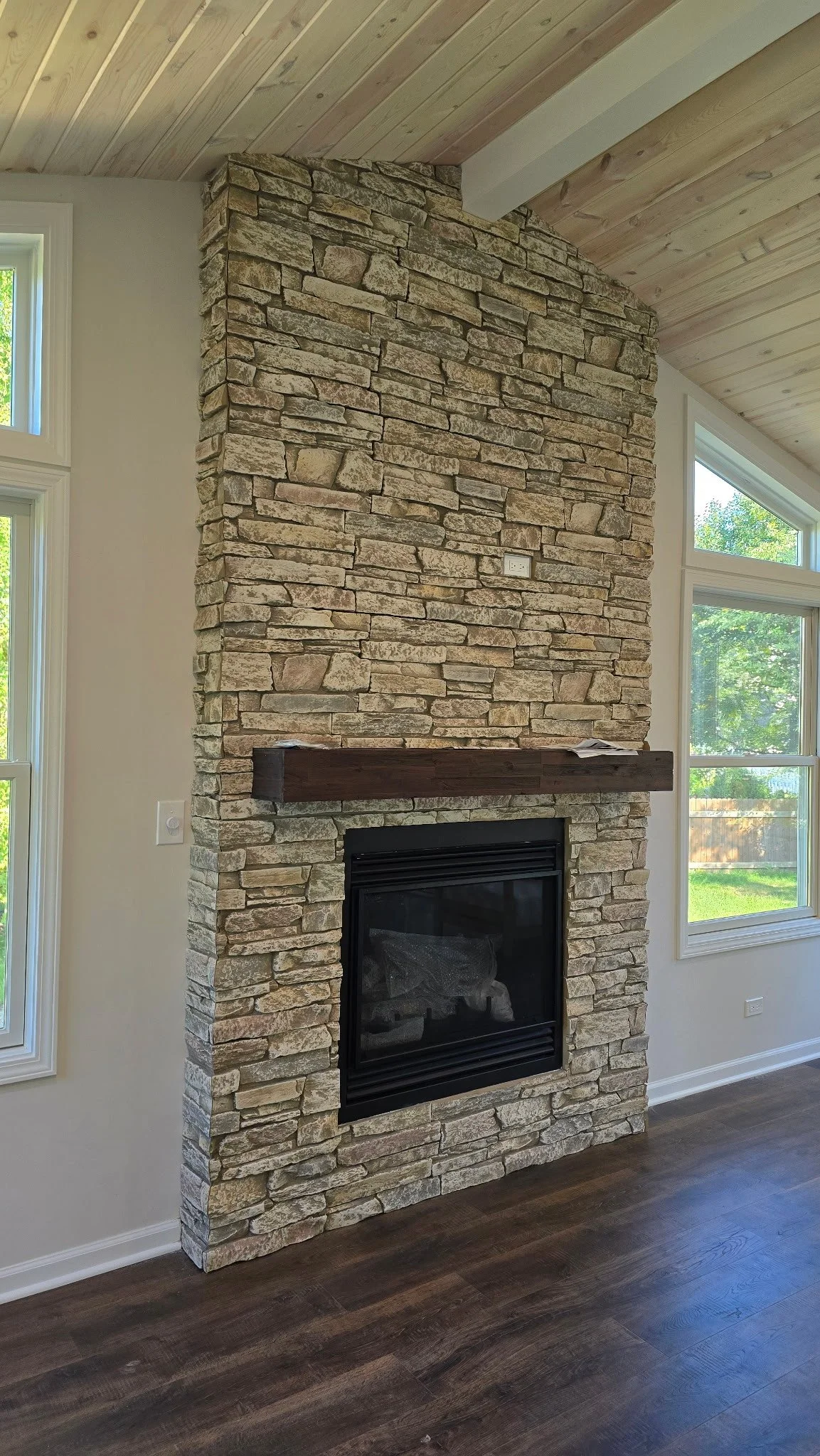 Stone fireplace with wooden mantel in a room with large windows and wood paneled ceiling.