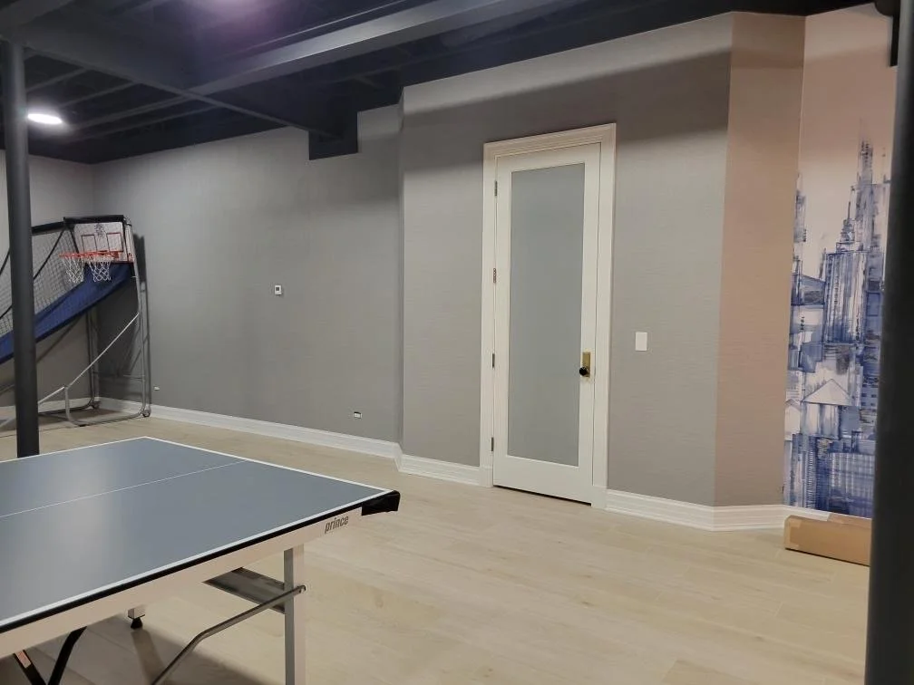An indoor recreational room with a ping pong table, a basketball arcade game, a door with frosted glass, and a cityscape mural on one wall.