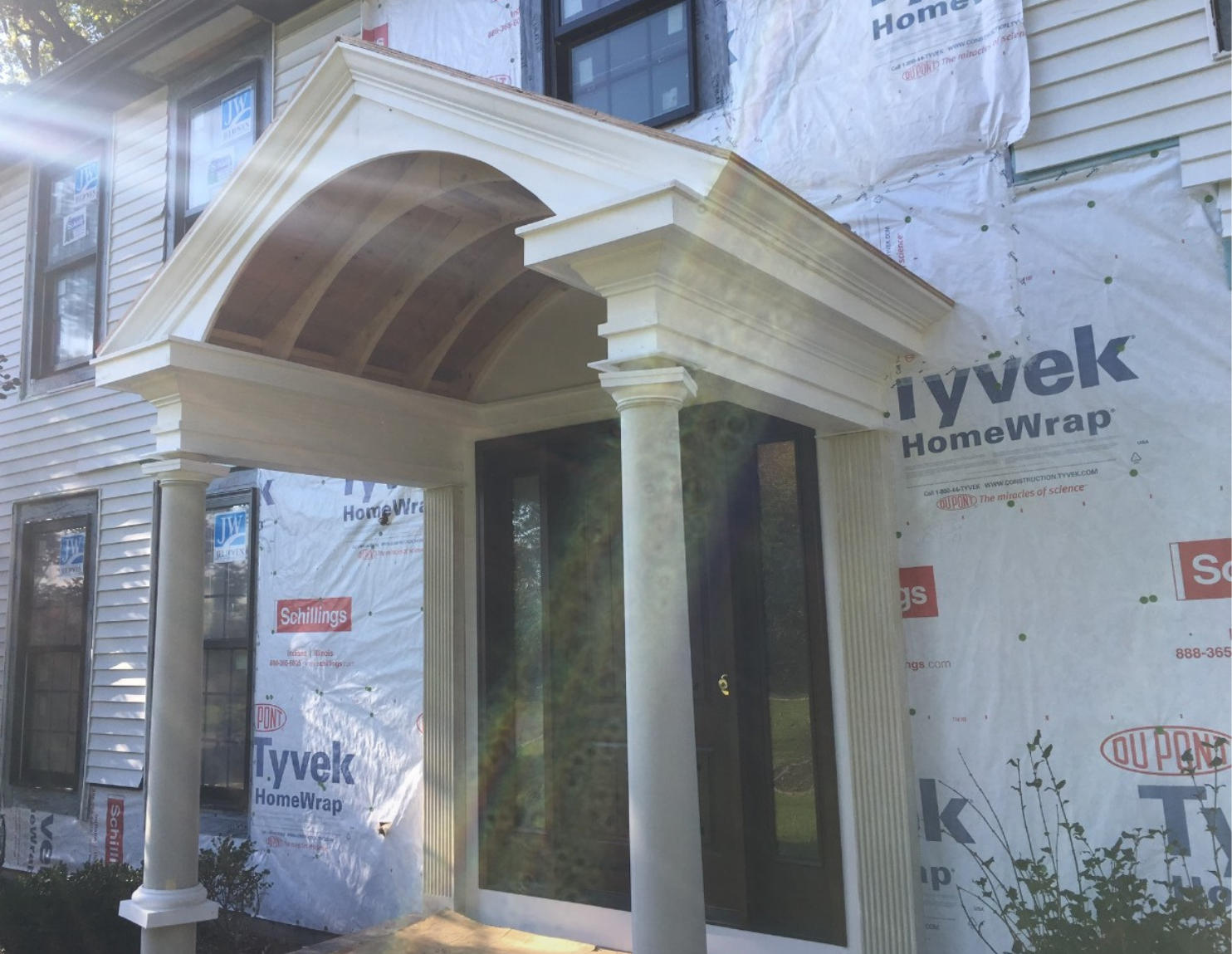 Front view of a house under construction, showing a white porch with columns, a glass door, and an arched pediment above the entrance, with partially installed siding and construction wrap.