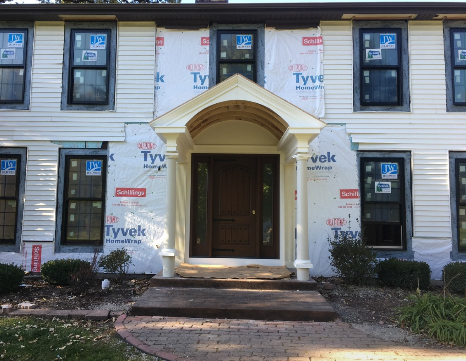 Front of a house under construction with siding partially installed, windows with labels, and a covered front porch with a wooden door and steps, surrounded by a brick pathway and landscaped garden beds.