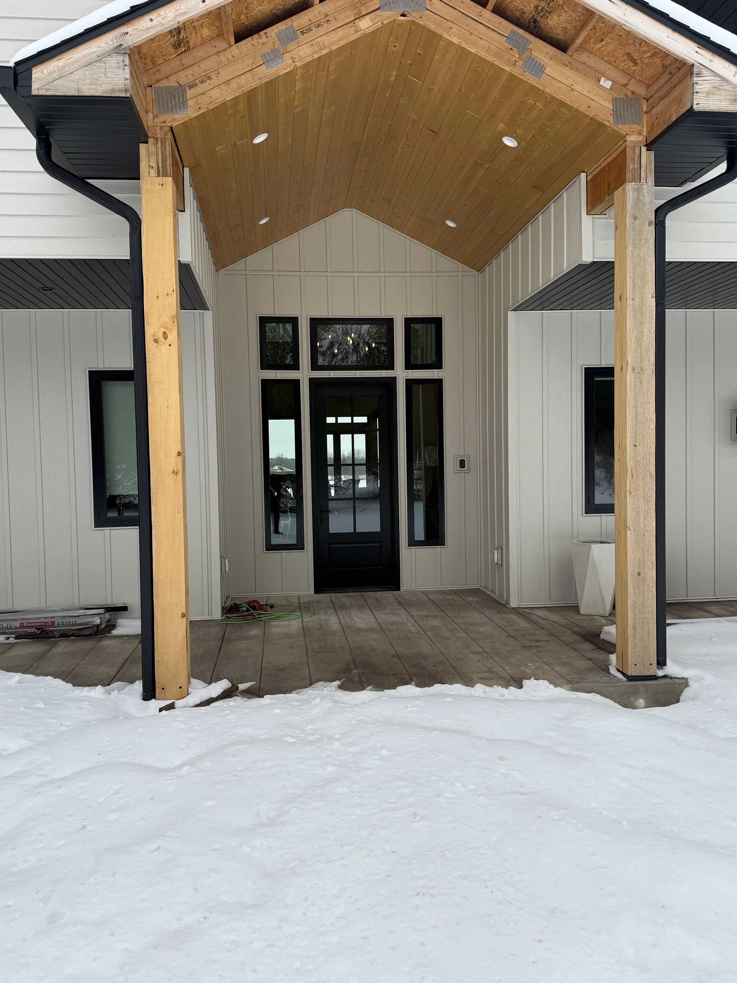 Front porch of a newly constructed house with snow on the ground, featuring a black door, large windows, and a wooden ceiling with recessed lights.