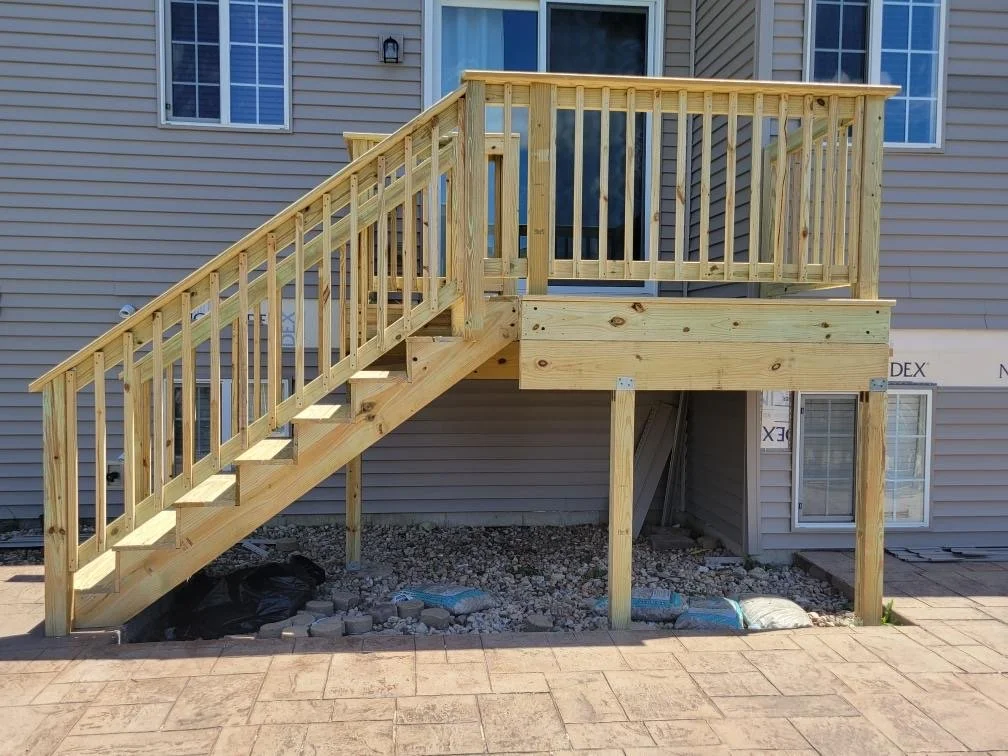 New wooden deck with stairs and railing attached to a house, under construction, with gravel and construction materials underneath.