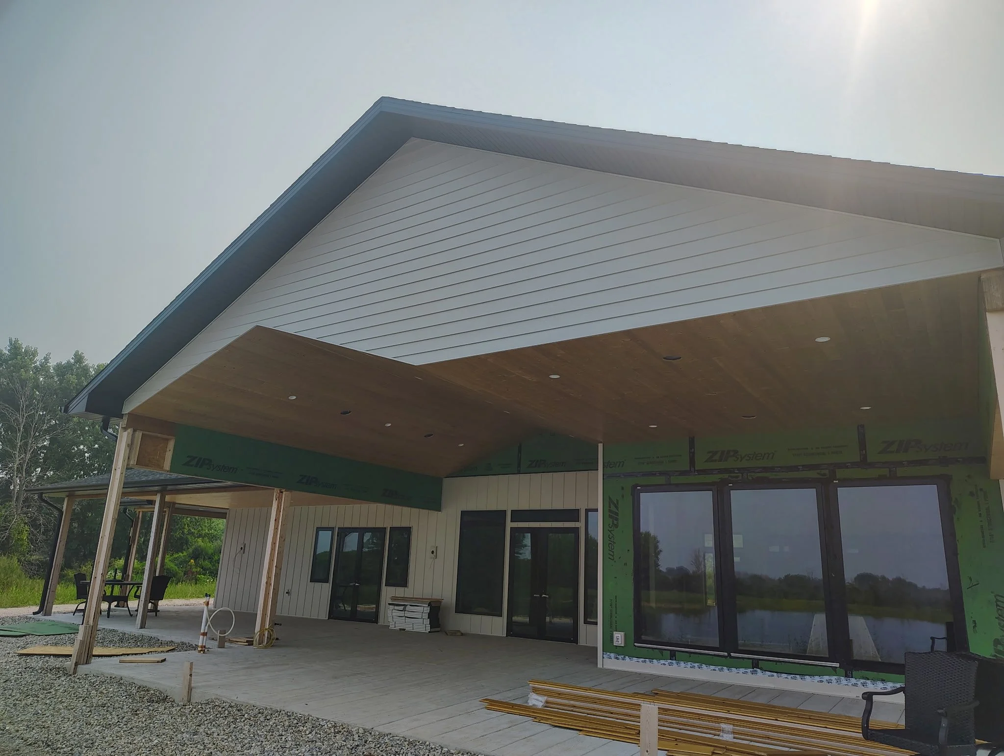 Under construction modern house with large windows, covered patio, and white siding, surrounded by greenery.