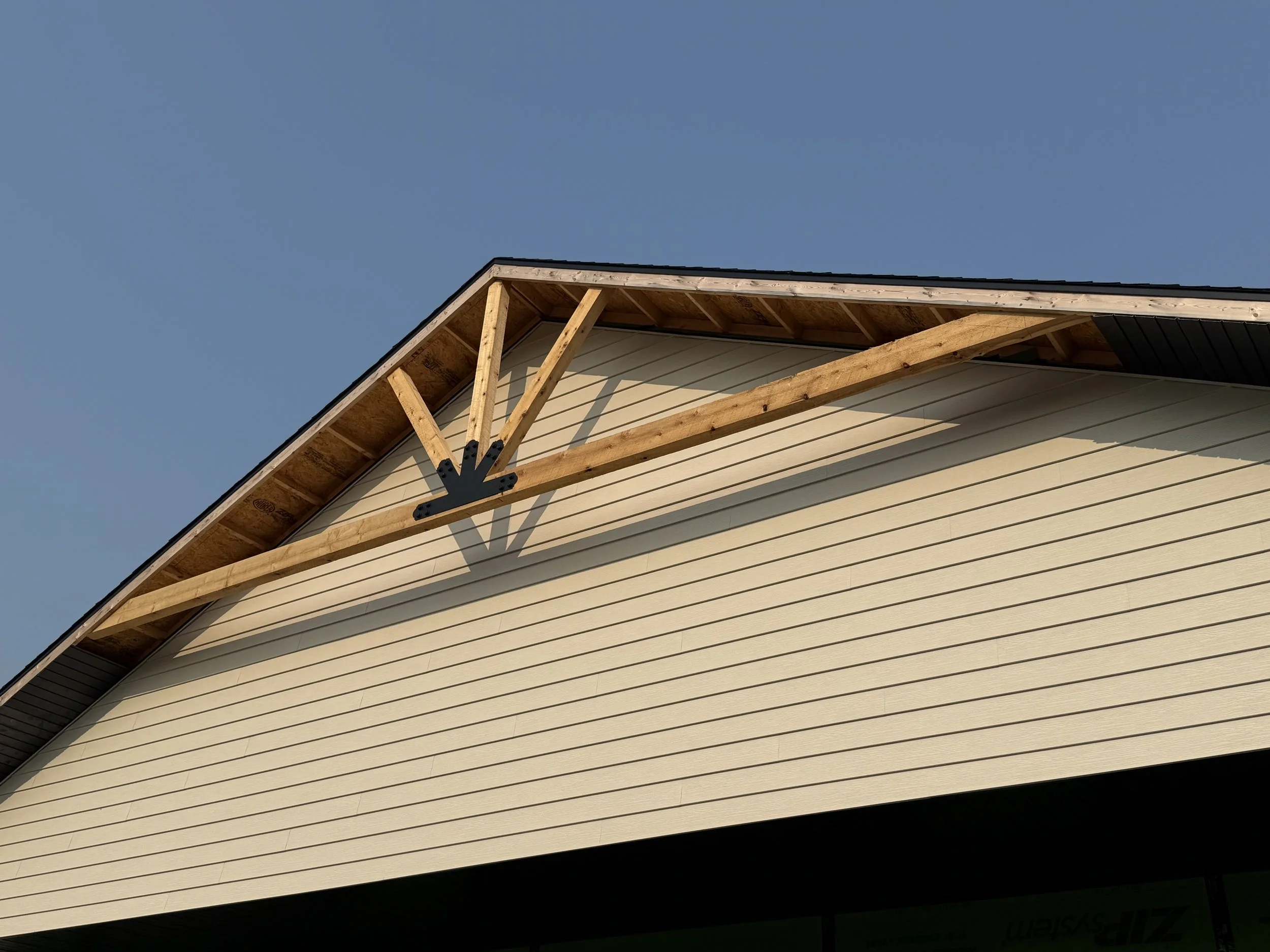 A house under construction showing the front gable with exposed wooden framework and beige horizontal siding, against a clear blue sky.