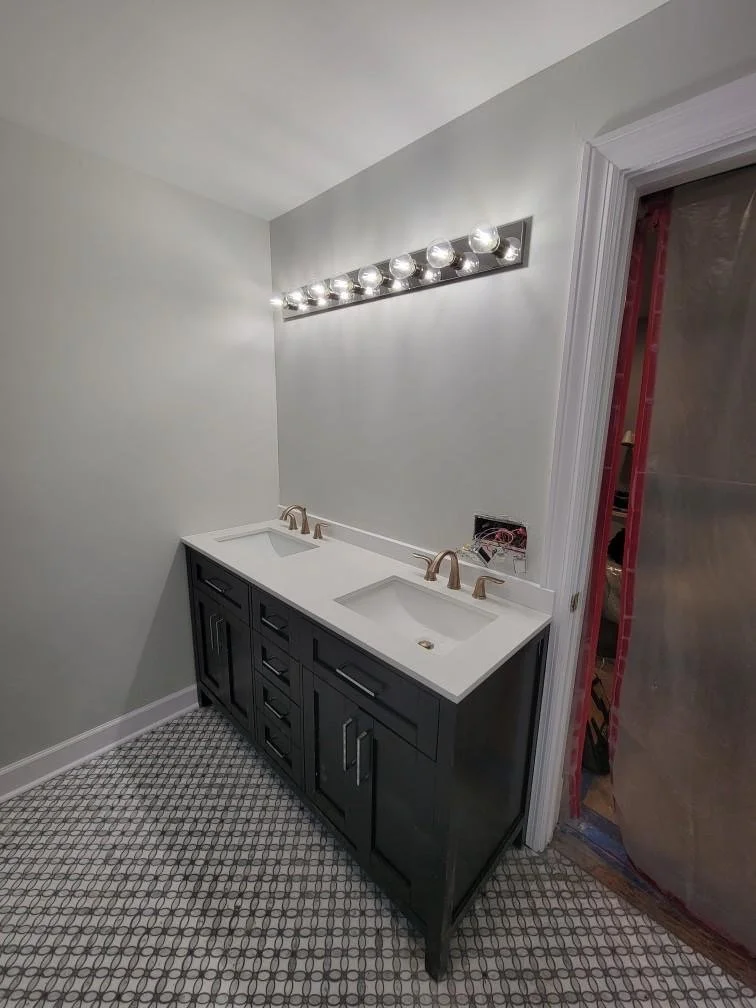 Bathroom vanity with two sinks, dark cabinetry, and modern fixtures; mirror and lighting above; bathroom under construction with covered doorway and patterned floor.