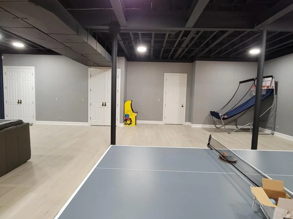 A basement recreational room with light wood flooring, gray walls, three closed white doors, sports equipment including a ping pong table, a basketball arcade game, and a shuffleboard court. The ceiling is unfinished with exposed beams and ducts.