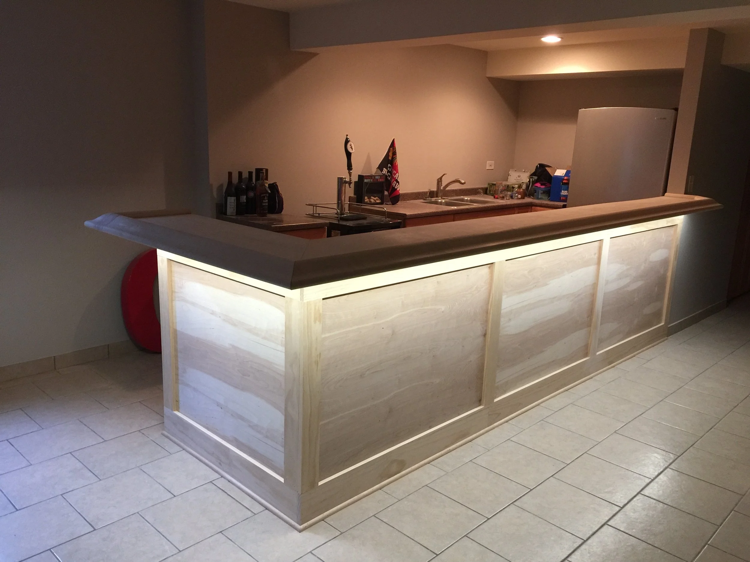A wooden bar counter with under-lighting in a room, with a kitchen area in the background containing bottles, a sink, and various kitchen items.