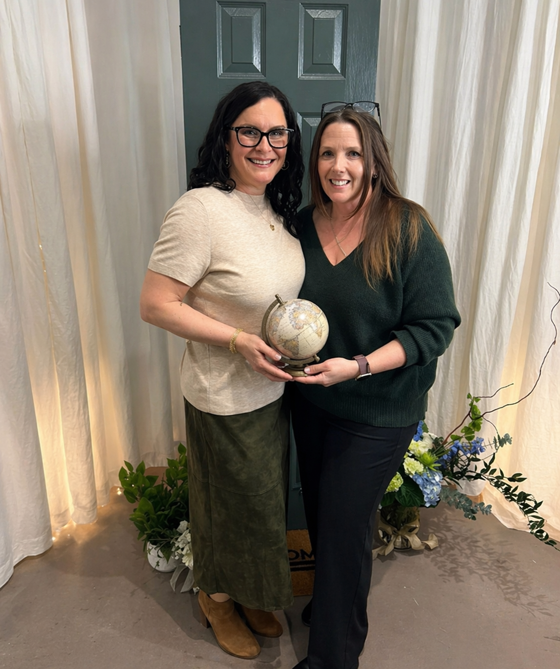Two women smiling and holding a small globe in a room with cream-colored curtains and floral arrangements.