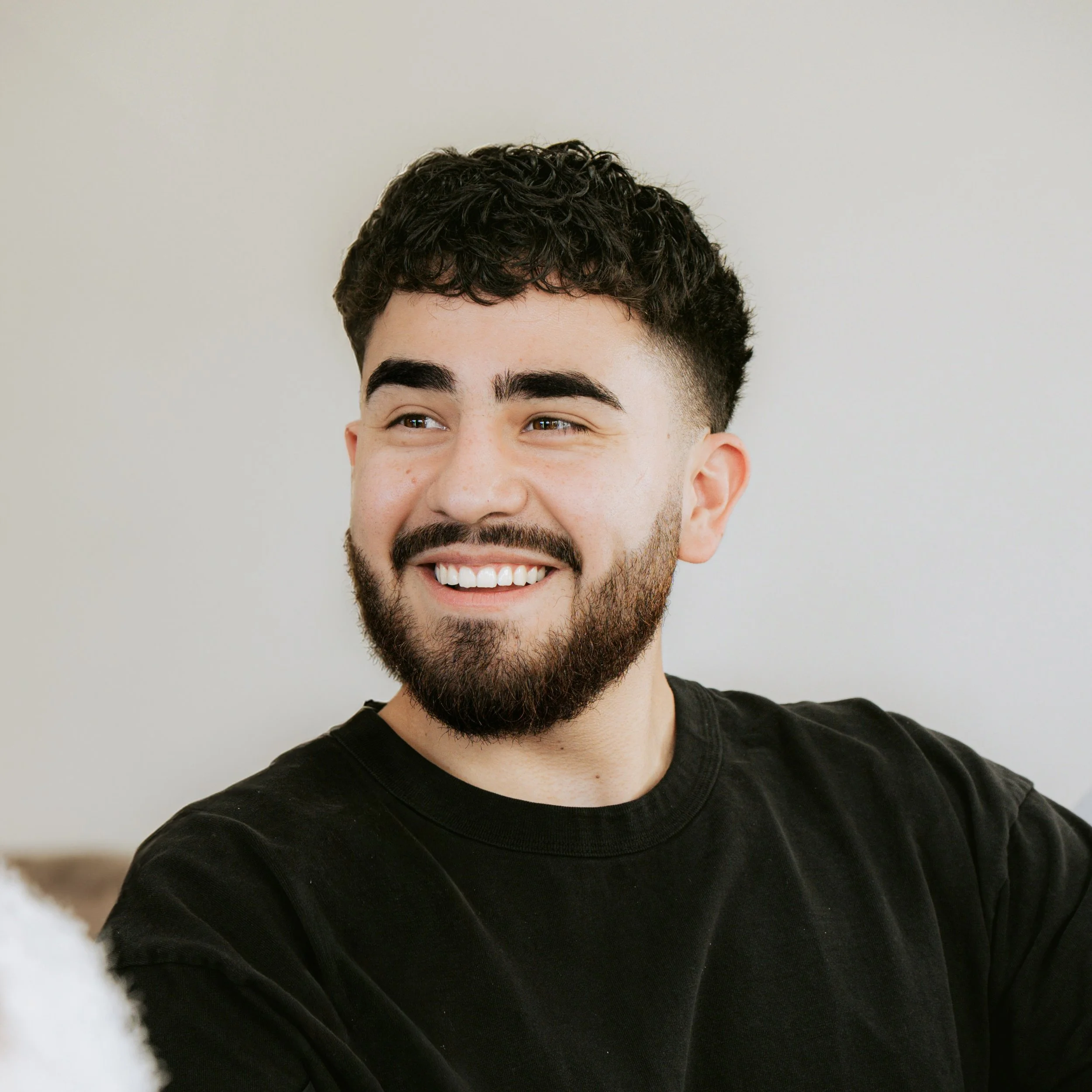 A smiling young man with dark curly hair, a beard, and wearing a black shirt.