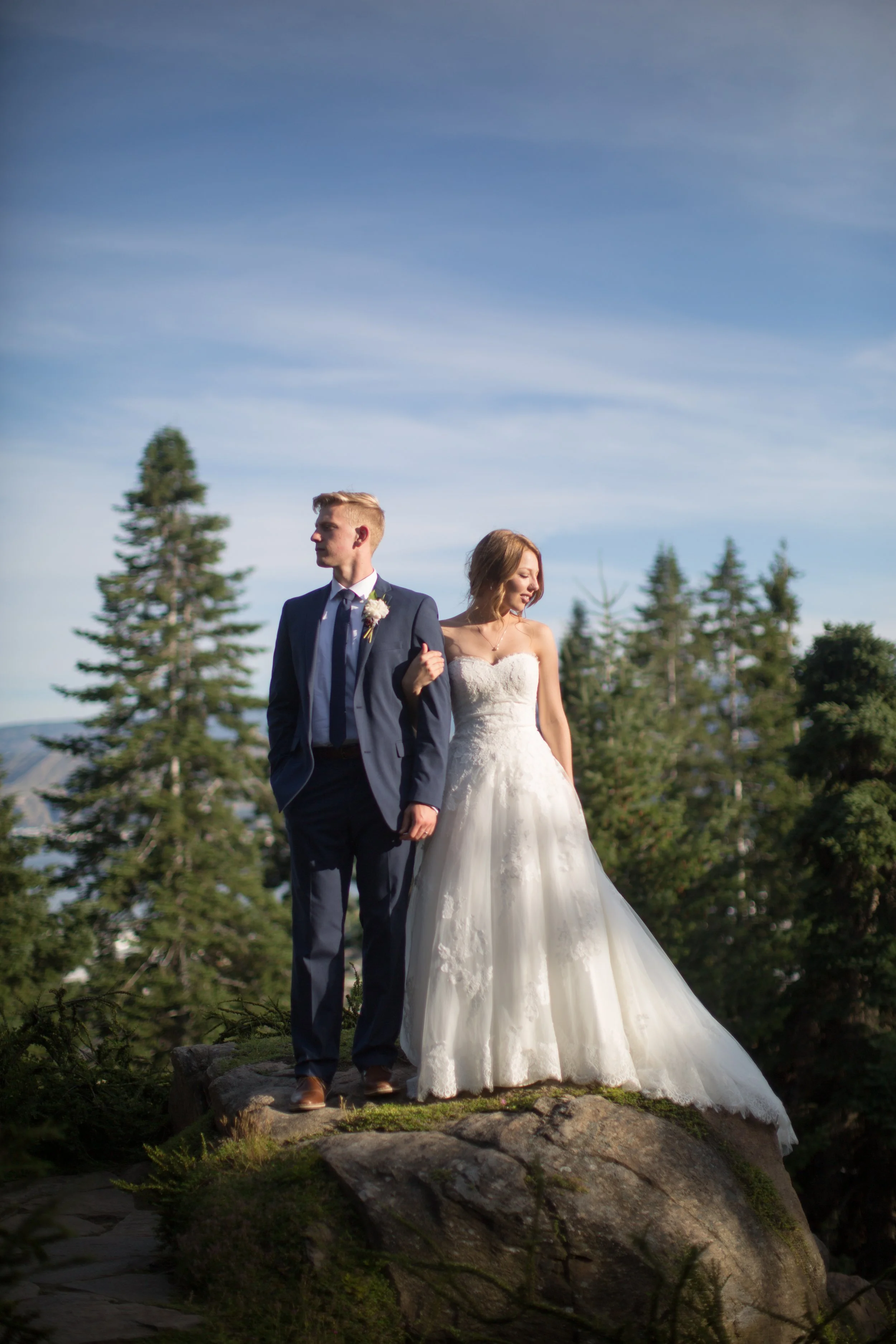 A bride and groom standing on a rock outdoors, surrounded by trees with a blue sky in the background during daytime.