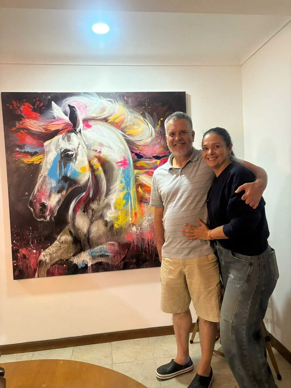 Una pareja sonriendo y abrazándose frente a una pintura de un caballo en un espacio de exhibición artística.