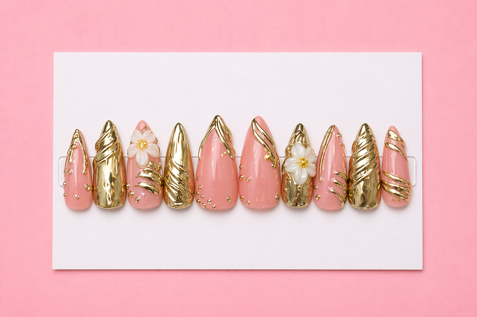 Set of decorative false nails with pink and gold designs, including floral accents and gold detailing, displayed on a white board with a pink background.