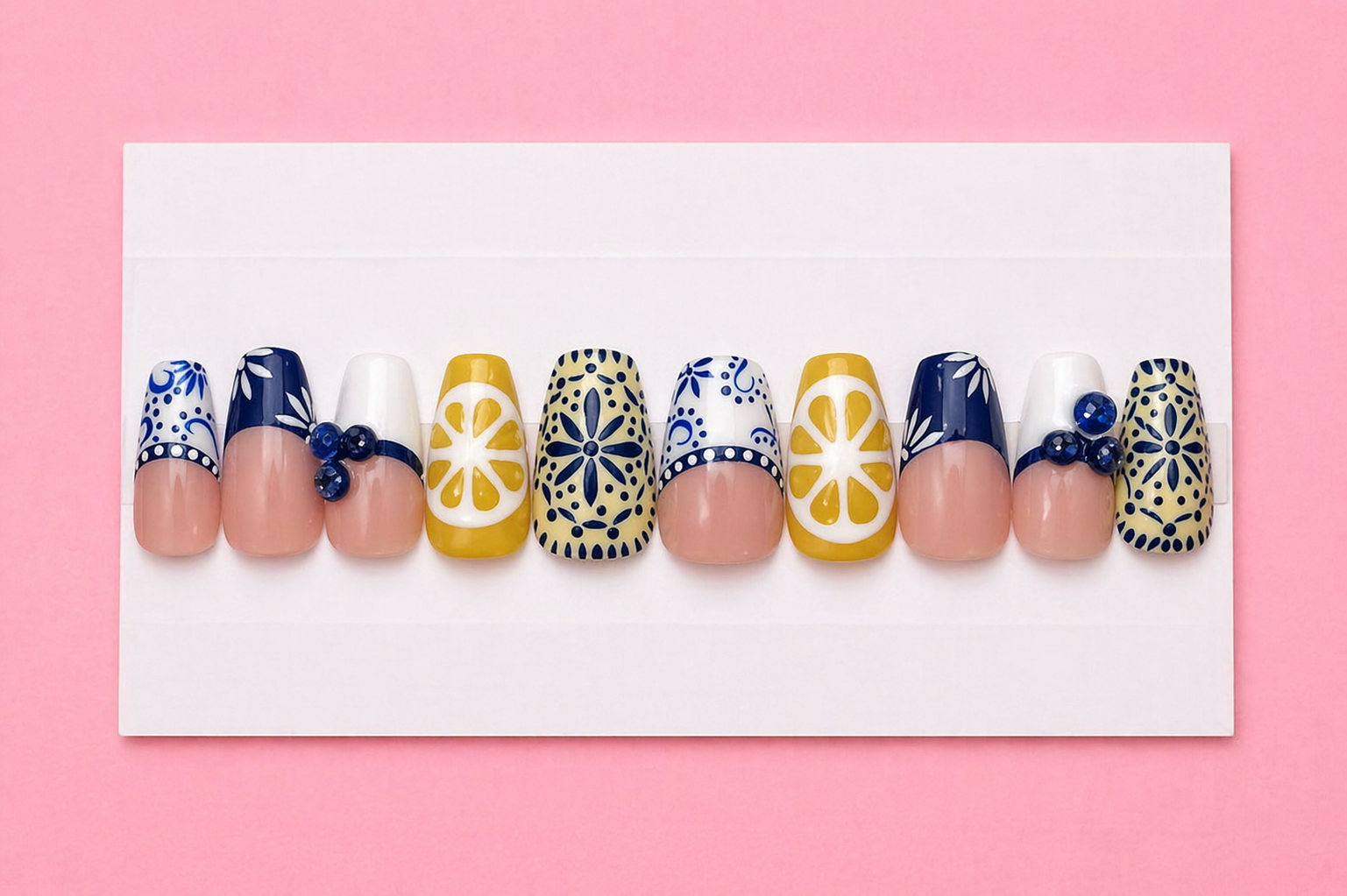 Set of decorative nail tips with blue, white, and yellow patterns, some with floral designs and blue gems, displayed on a white board against a pink background.