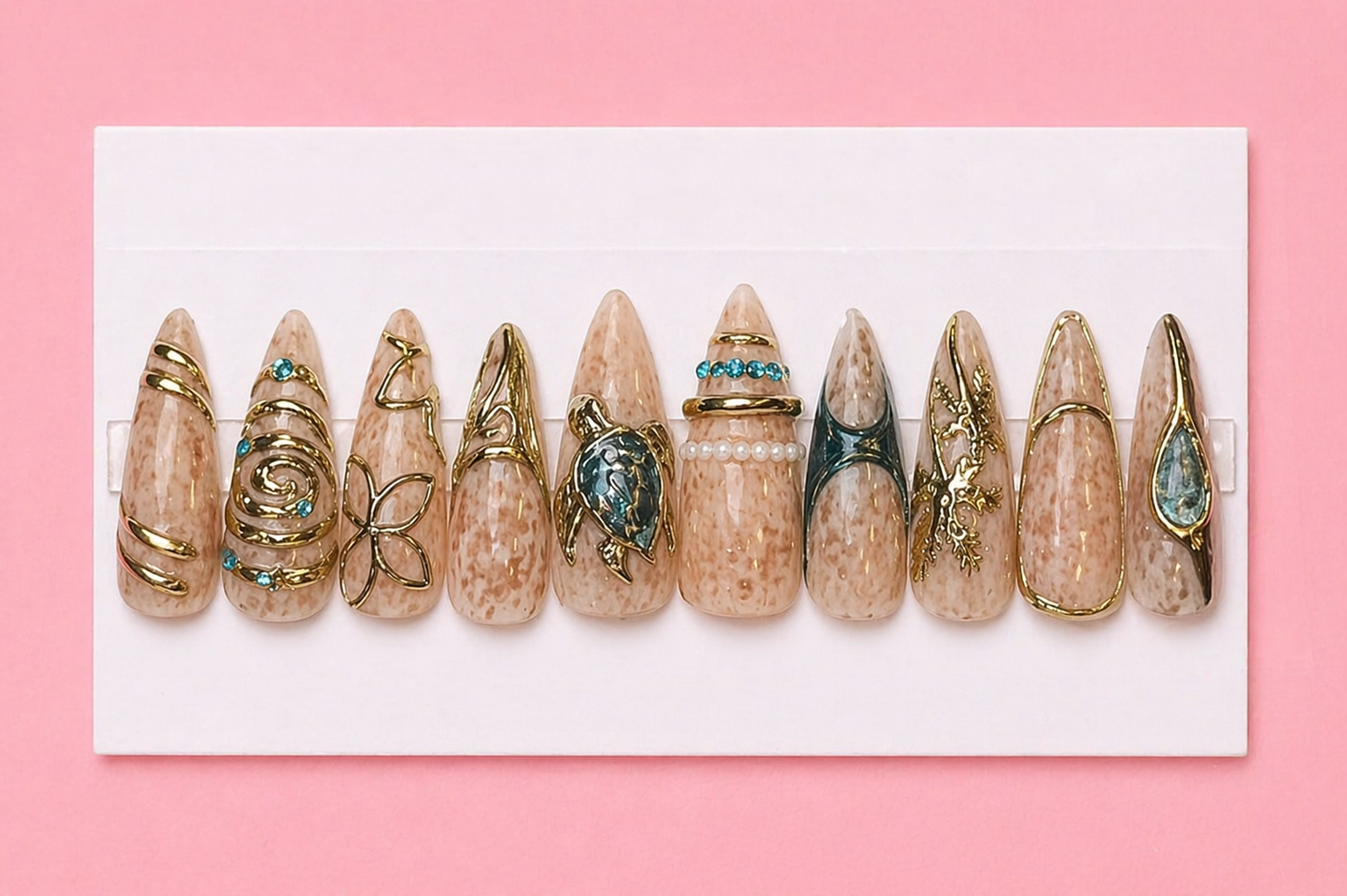A display of artificially decorated nails with various gold and gemstone jewelry attachments, set against a pink background.