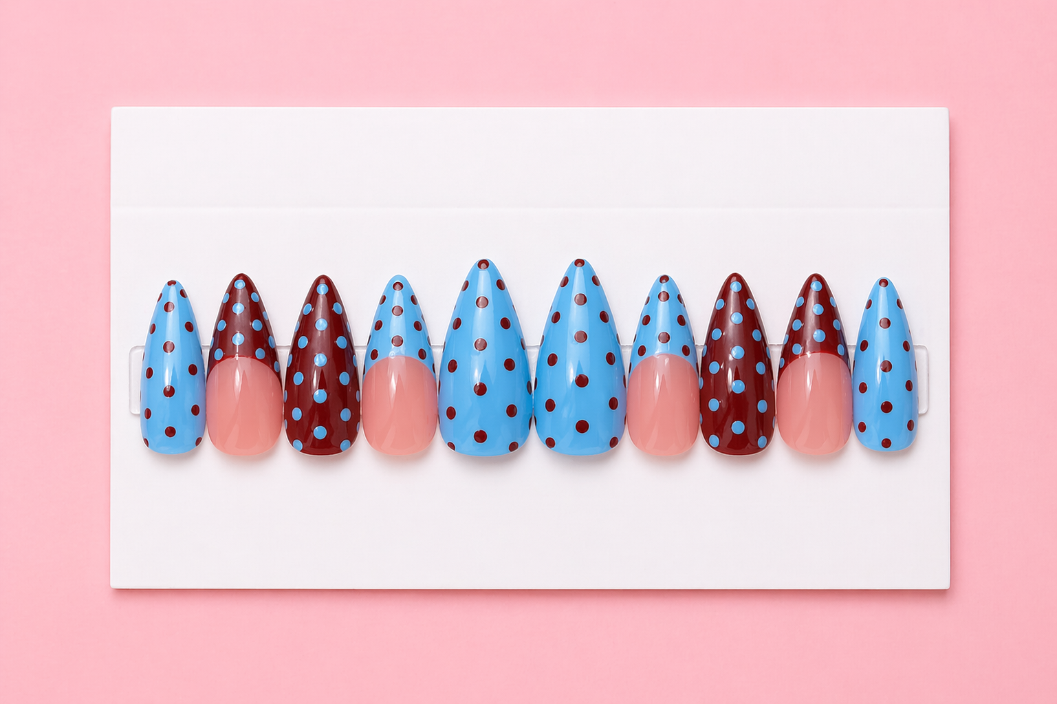 A set of ten false nails with blue and red polka dot designs arranged on a white tray against a pink background.