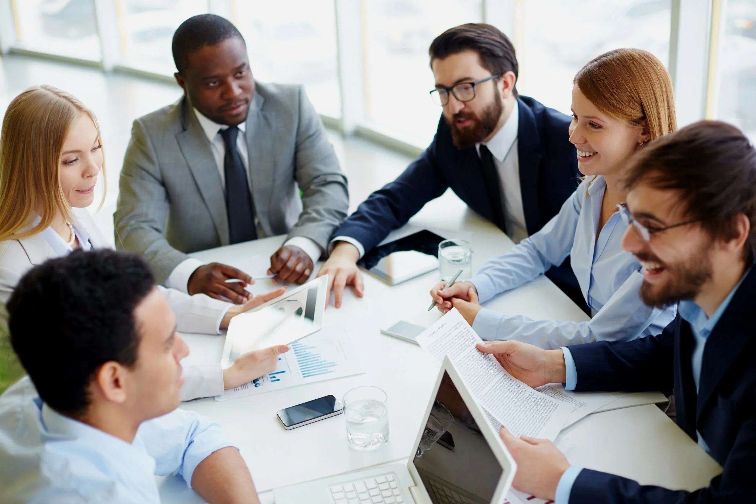 A group of six diverse professionals in business attire having a meeting in a conference room with large windows, discussing documents and charts.