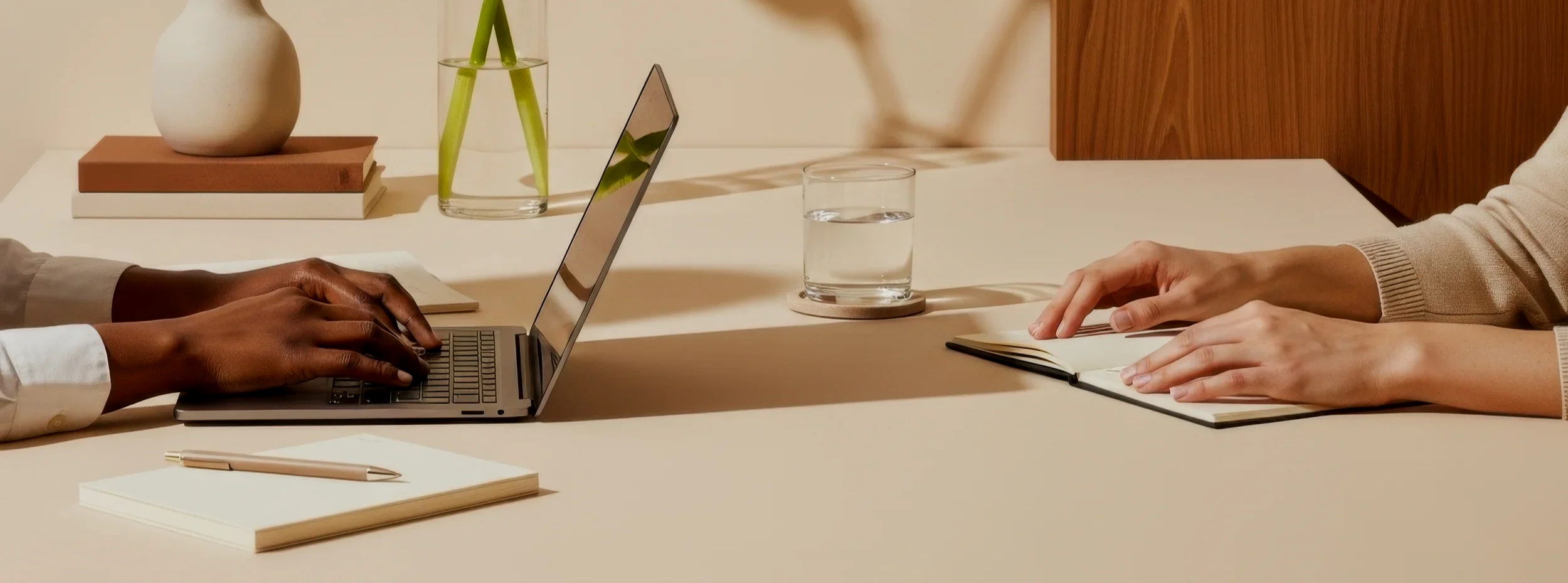 Two people are sitting at a table, one using a laptop and the other writing in a notebook, with a glass of water and decorative items on the table.