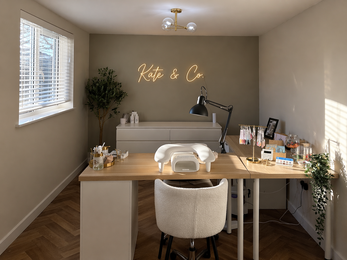 A modern nail salon workspace with a neon sign that says 'Kate & Co.' on the wall. The desk has various manicure tools, nail polish bottles, a lamp. There are two windows with blinds, a chair, a potted plant, and a cabinet for supplies.