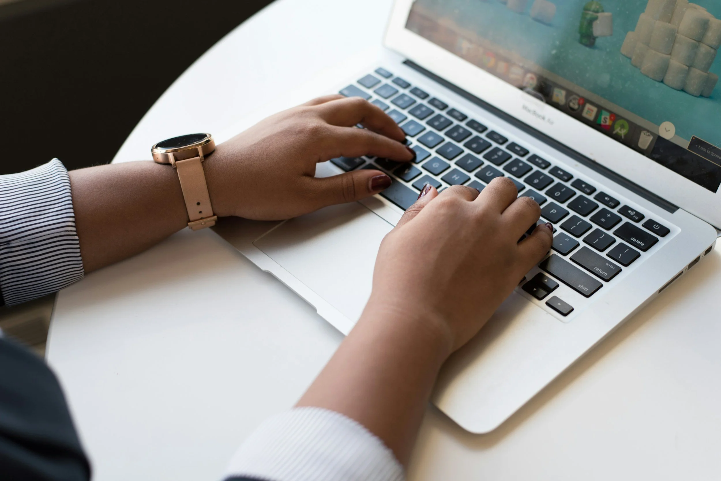 A person wearing a black and white striped shirt and a rose gold smartwatch is typing on a silver laptop with a colorful game open on the screen, sitting at a white table.