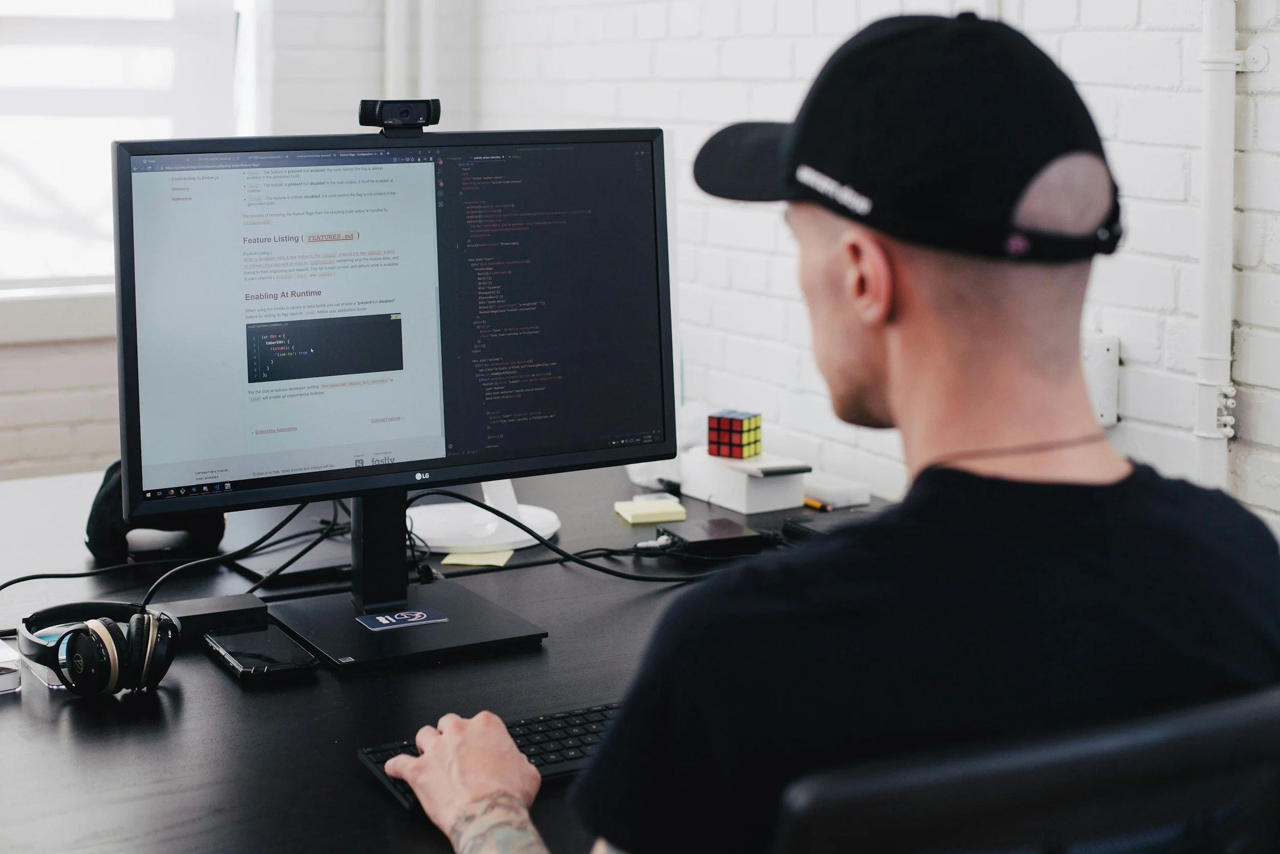A person with a tattoo on their arm wearing a black cap working at a computer with dual monitors in a white-brick room. The monitors display code and a webpage. On the desk, there are headphones, a Rubik's cube, and a smartphone.